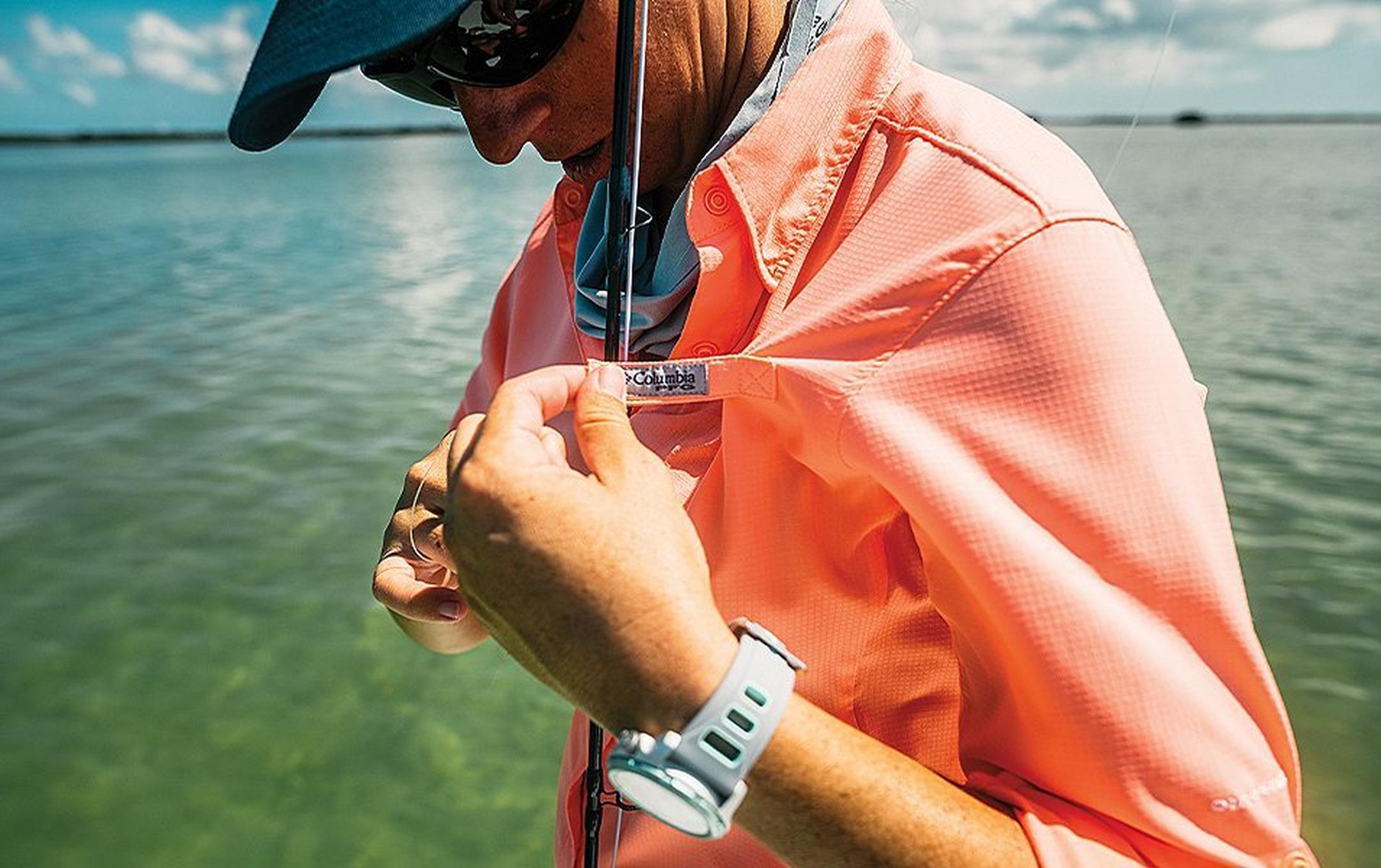 A woman in an orange Columbia Sportswear PFG shirt holds a fishing rod while standing by a river.