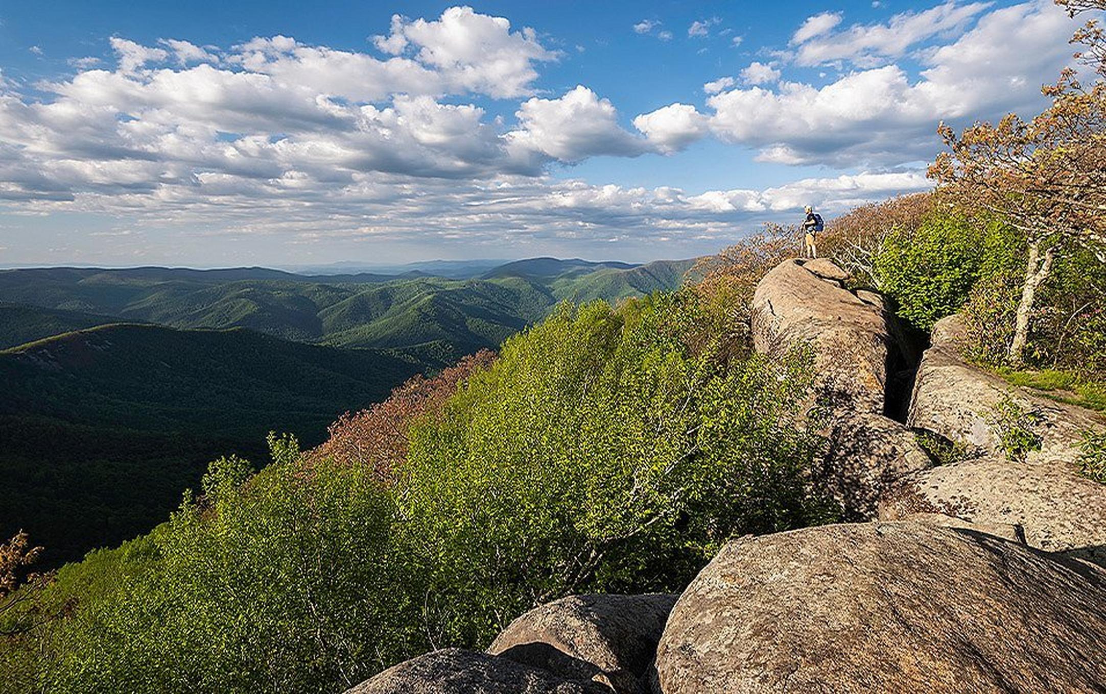 A hiker stands on a large boulder, gazing at rolling ridges and valleys in the distance.