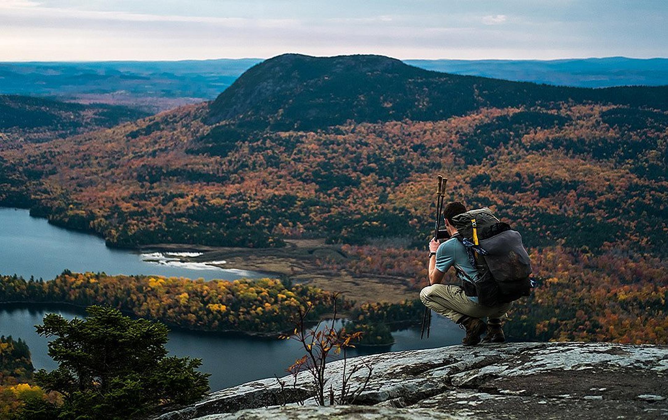 A hiker with a big backpack and hiking poles crouches on a rocky ridge, photographing a lake and tree-covered knoll.