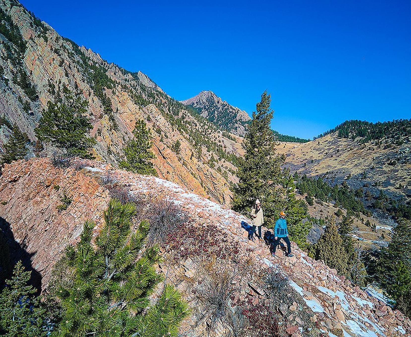 A pair of hikers walk down a steep, rocky trail in a beautiful mountainous setting.