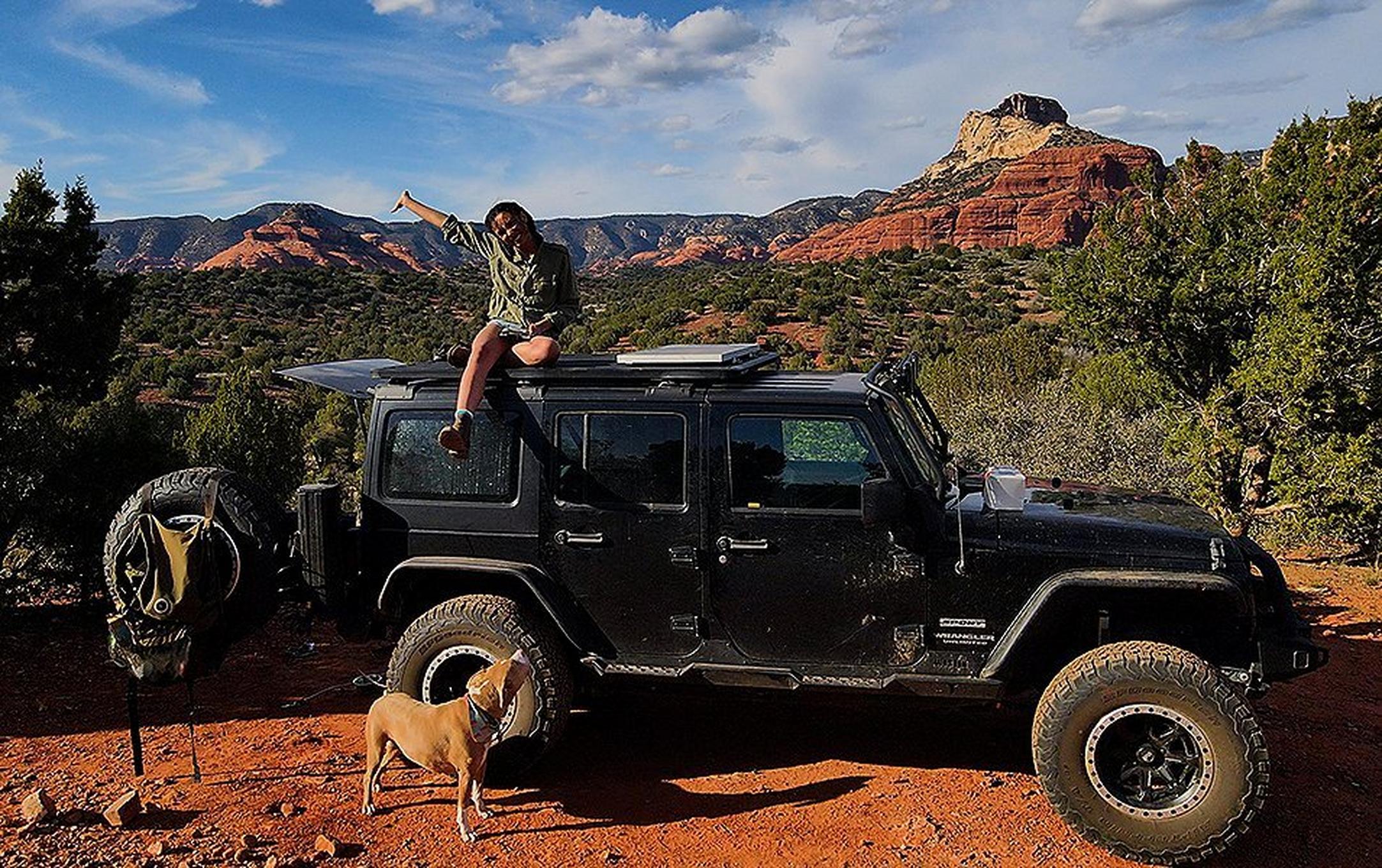 A woman on an overlanding adventure sits on top of a Jeep in the high desert smiling with her arm in the air as her dog looks up at her.