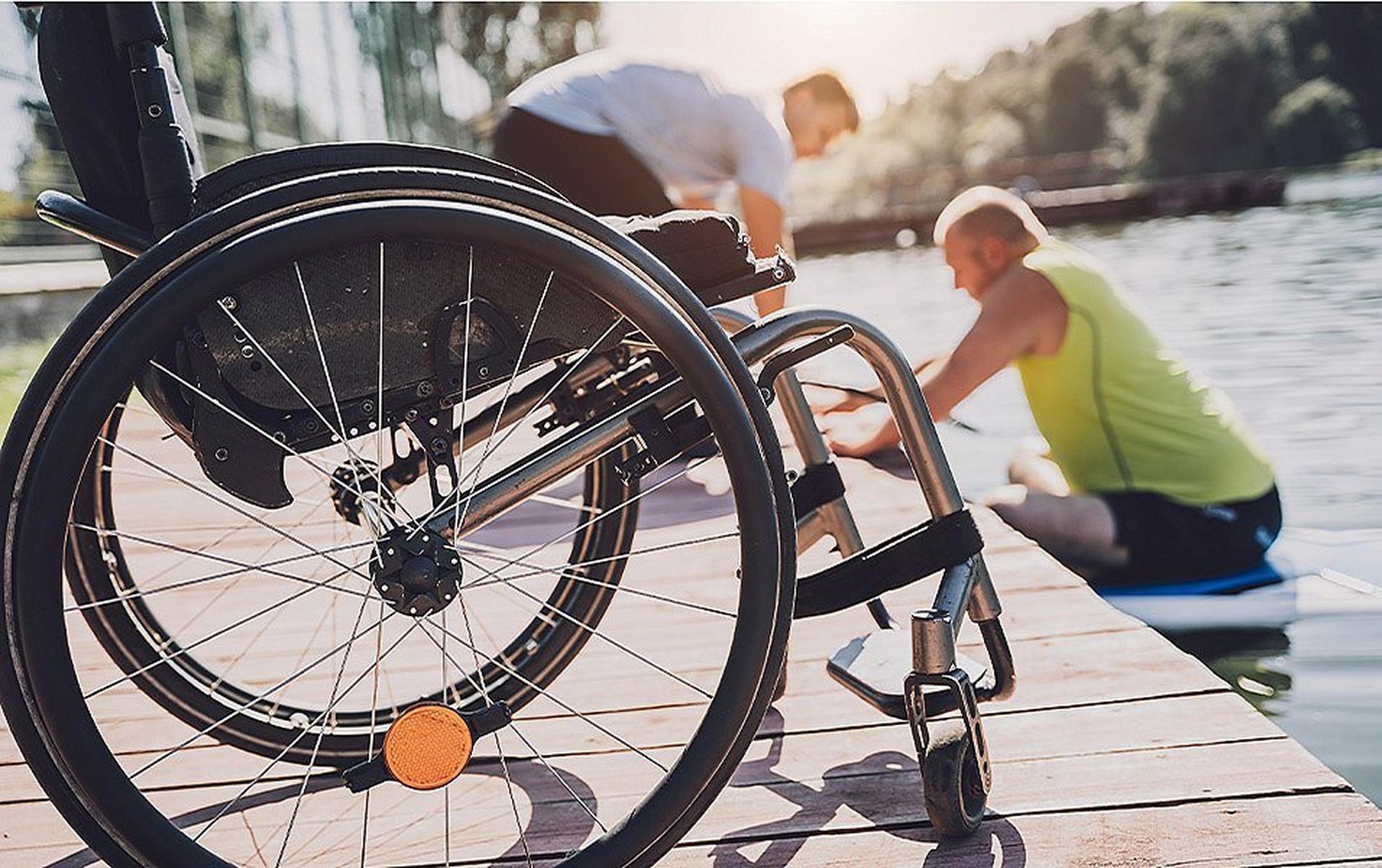 With a wheelchair in the foreground, a man with a lower body disability is sitting on a paddleboard. Another man is assisting him.