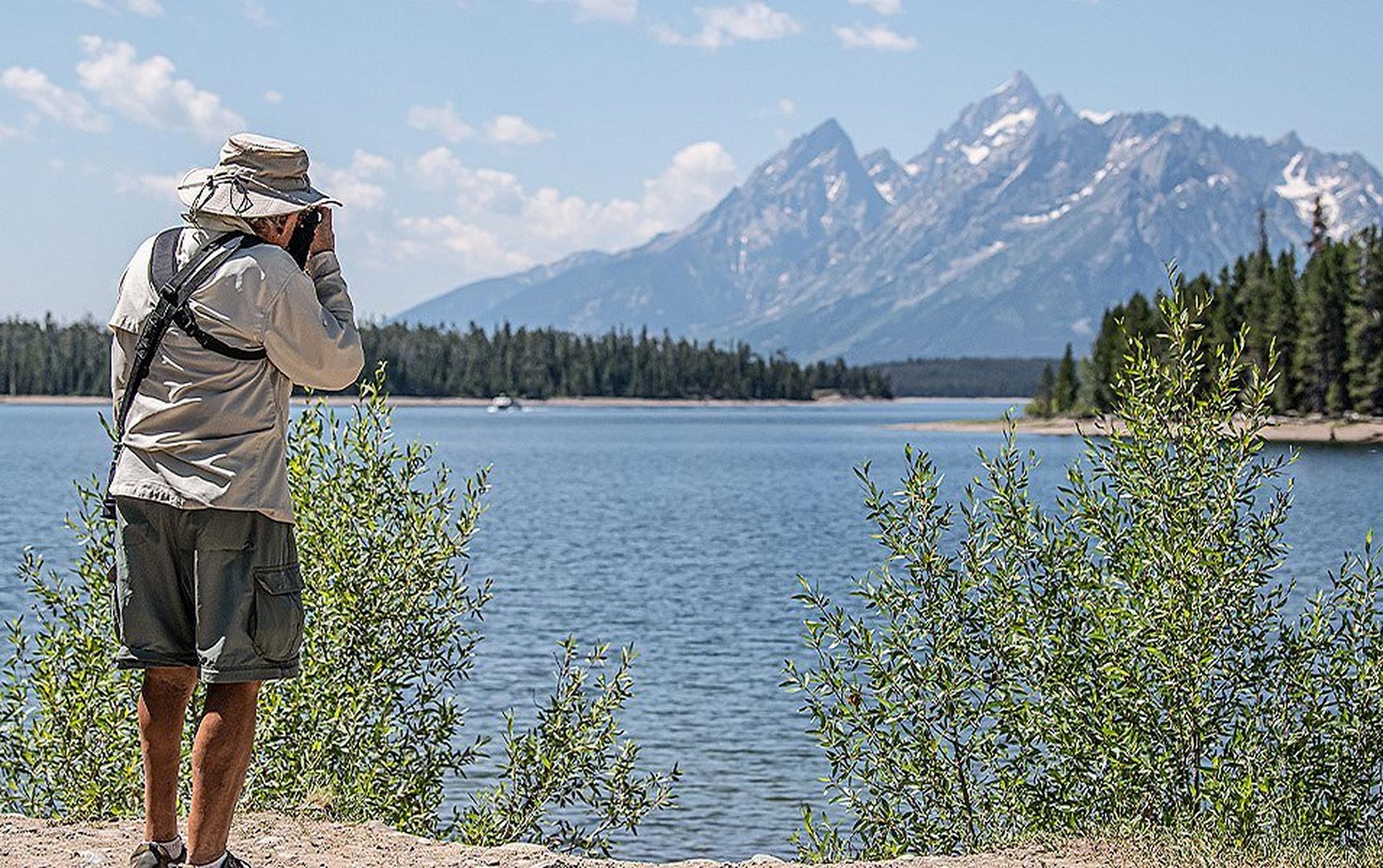 A man wearing shorts and a bucket hat is standing on a lakeside path. He is taking a photo of a mountain range in the distance.