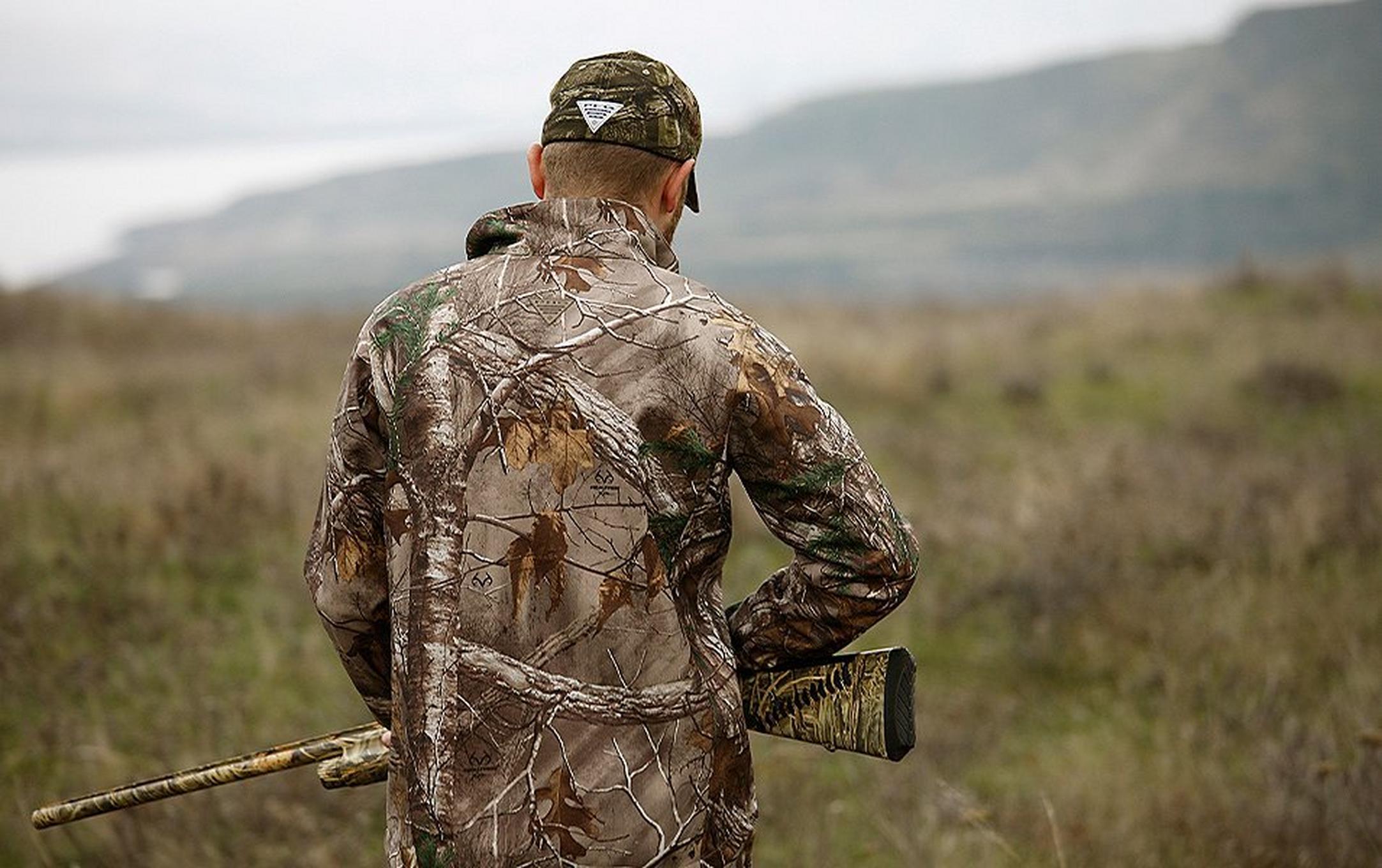 A hunter stands in a field holding a rifle with his back to the camera wearing Columbia PHG clothing.