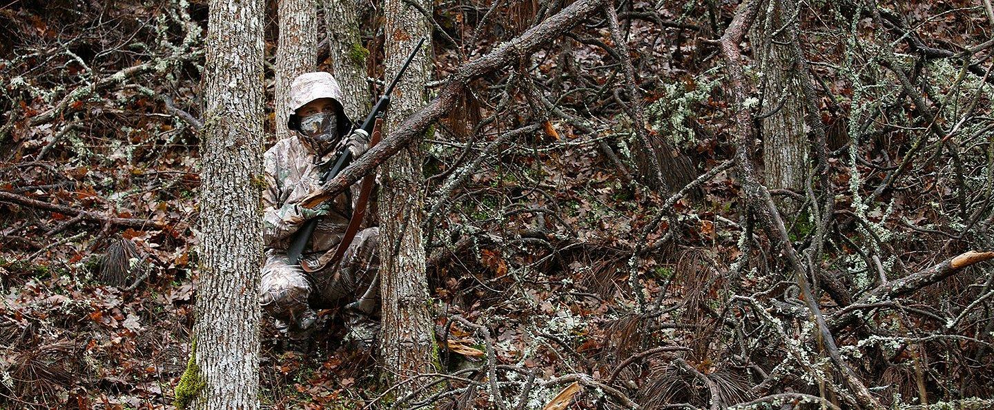 A hunter crouches behind a tree wearing full-body camouflage in the woods.