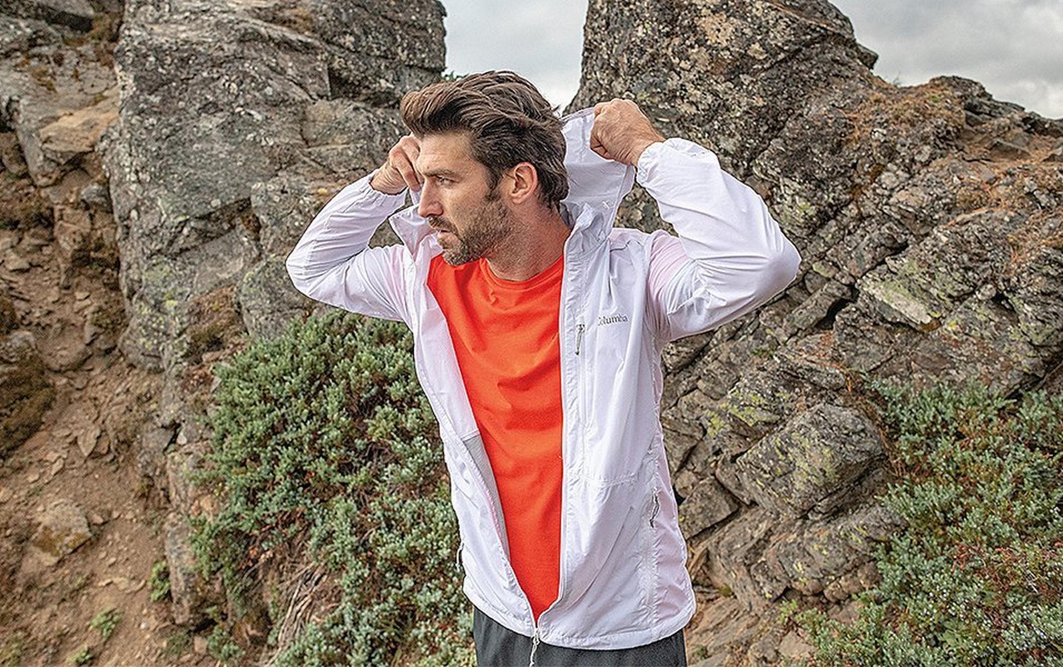 A man fixes the hood of his windbreaker jacket while standing in front of a jagged rock outcrop.