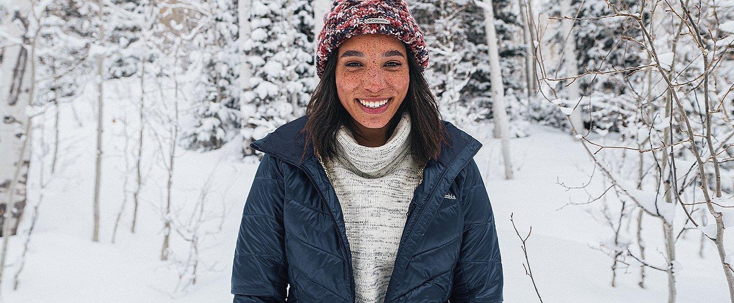 A woman is standing in the foreground of a snow-covered forest wearing a puffer jacket.