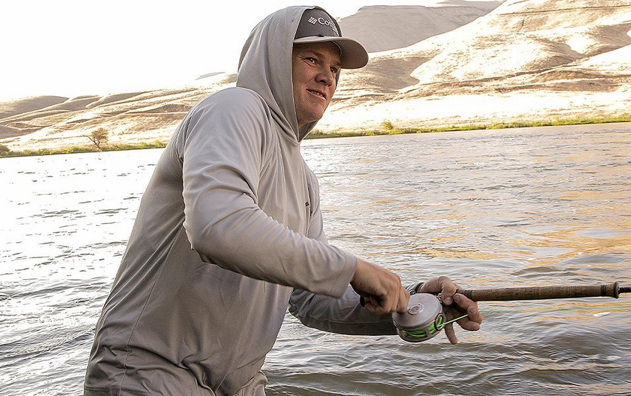 Pro angler Curtis Ciszek stands in a river fishing with high desert plains in the background.