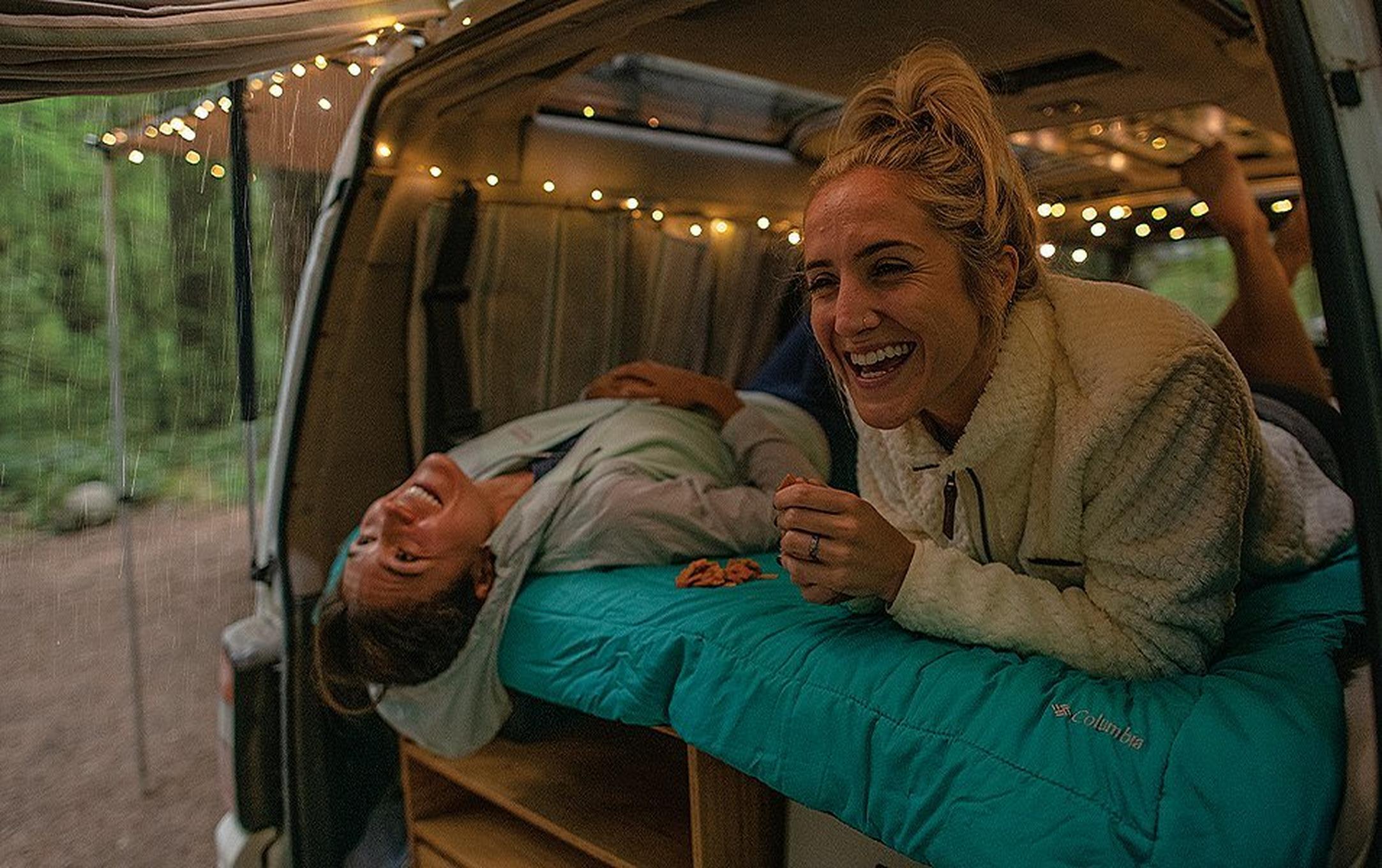 A woman in a white fleece jacket laughs in the back of a camper van while another woman lies next to her upside down as she looks up and smiles.