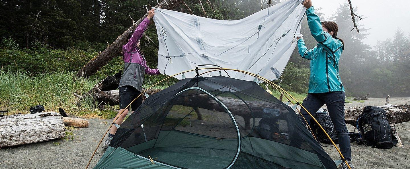 Two women hang a tarp over a tent while camping in the rain.