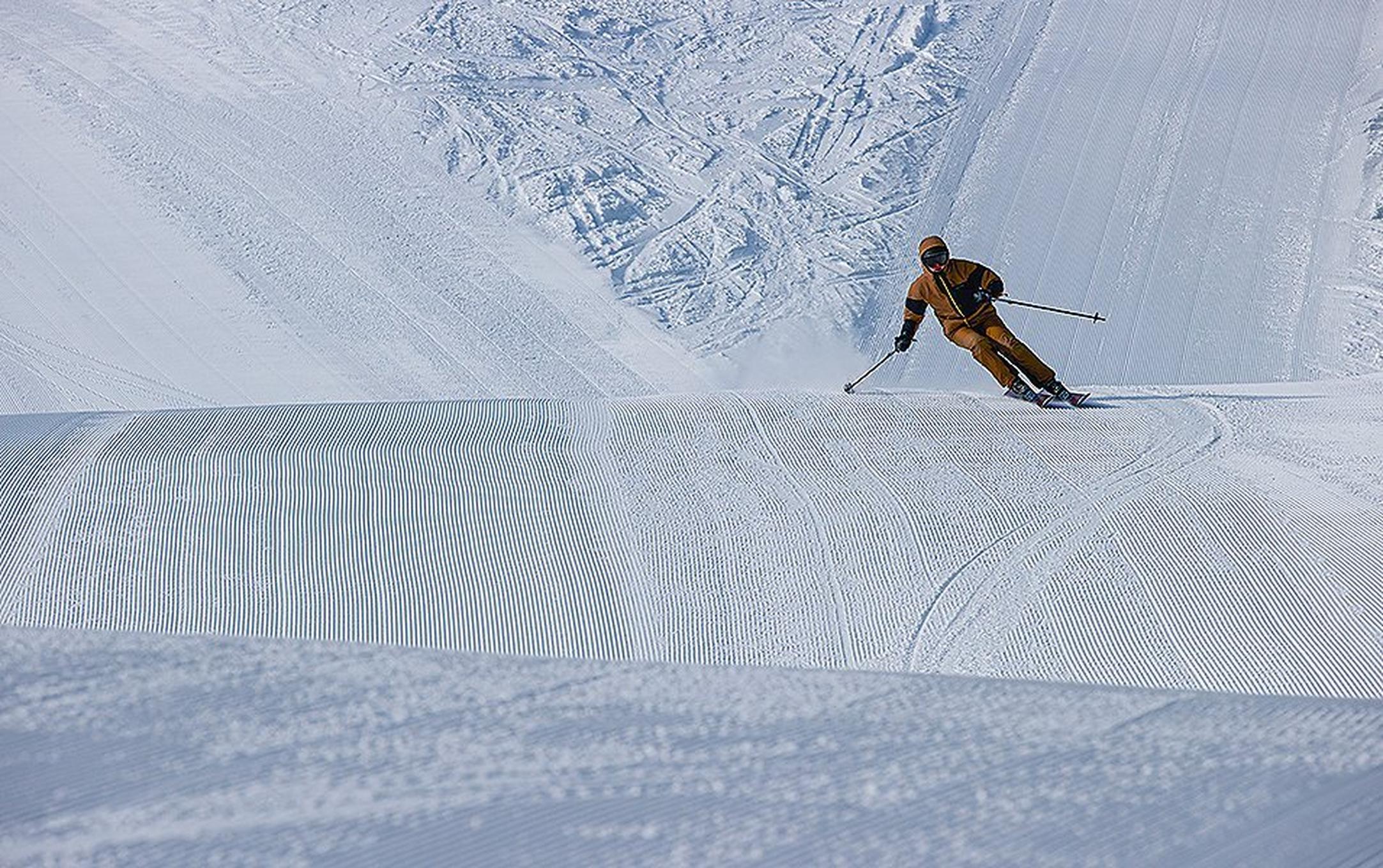 A man in an orange jacket skis down a patch of corduroy on a freshly groomed ski slope.