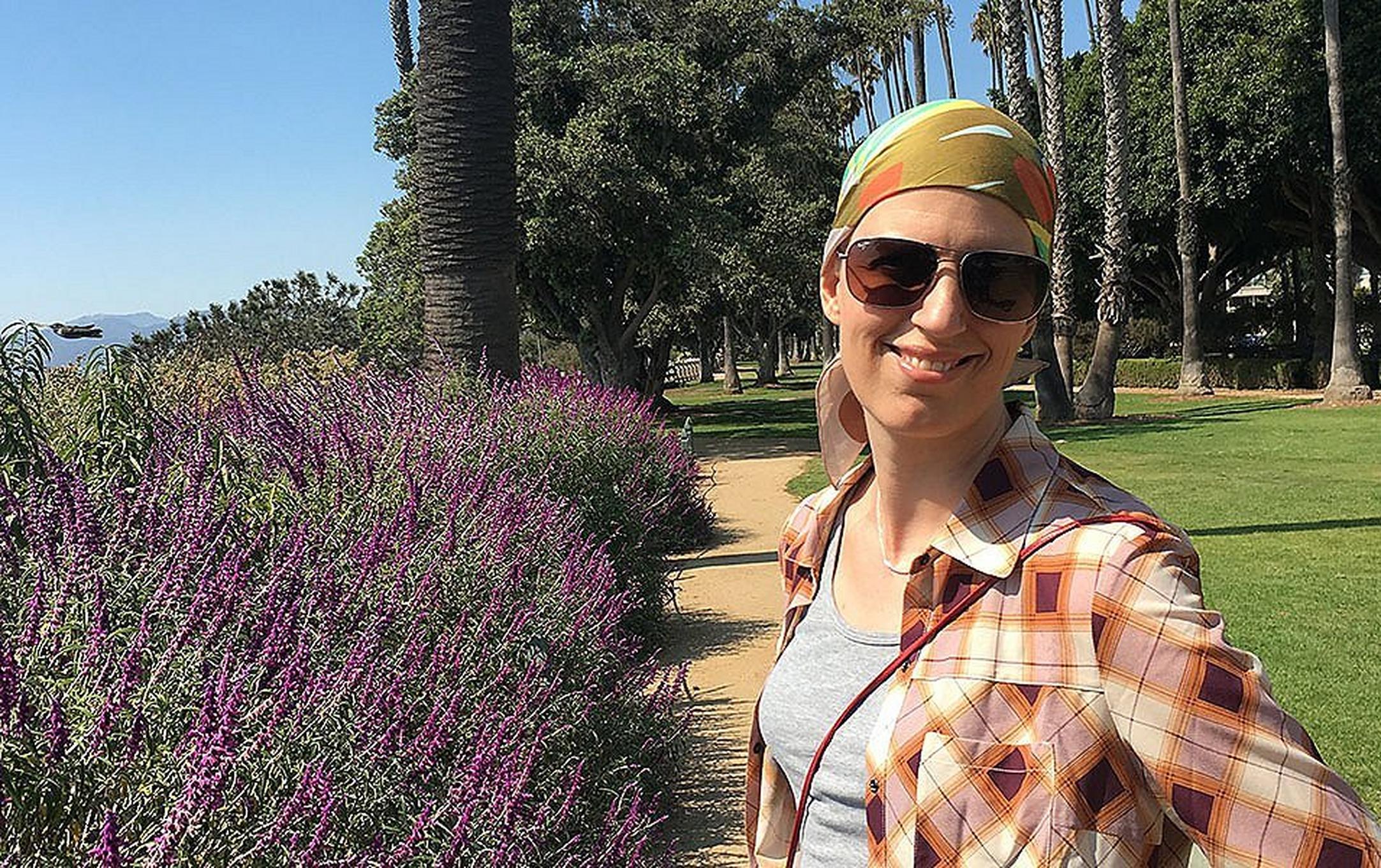 A woman in a plaid shirt and headscarf smiles at the camera on a sunny day at Santa Monica’s Palisades Park.