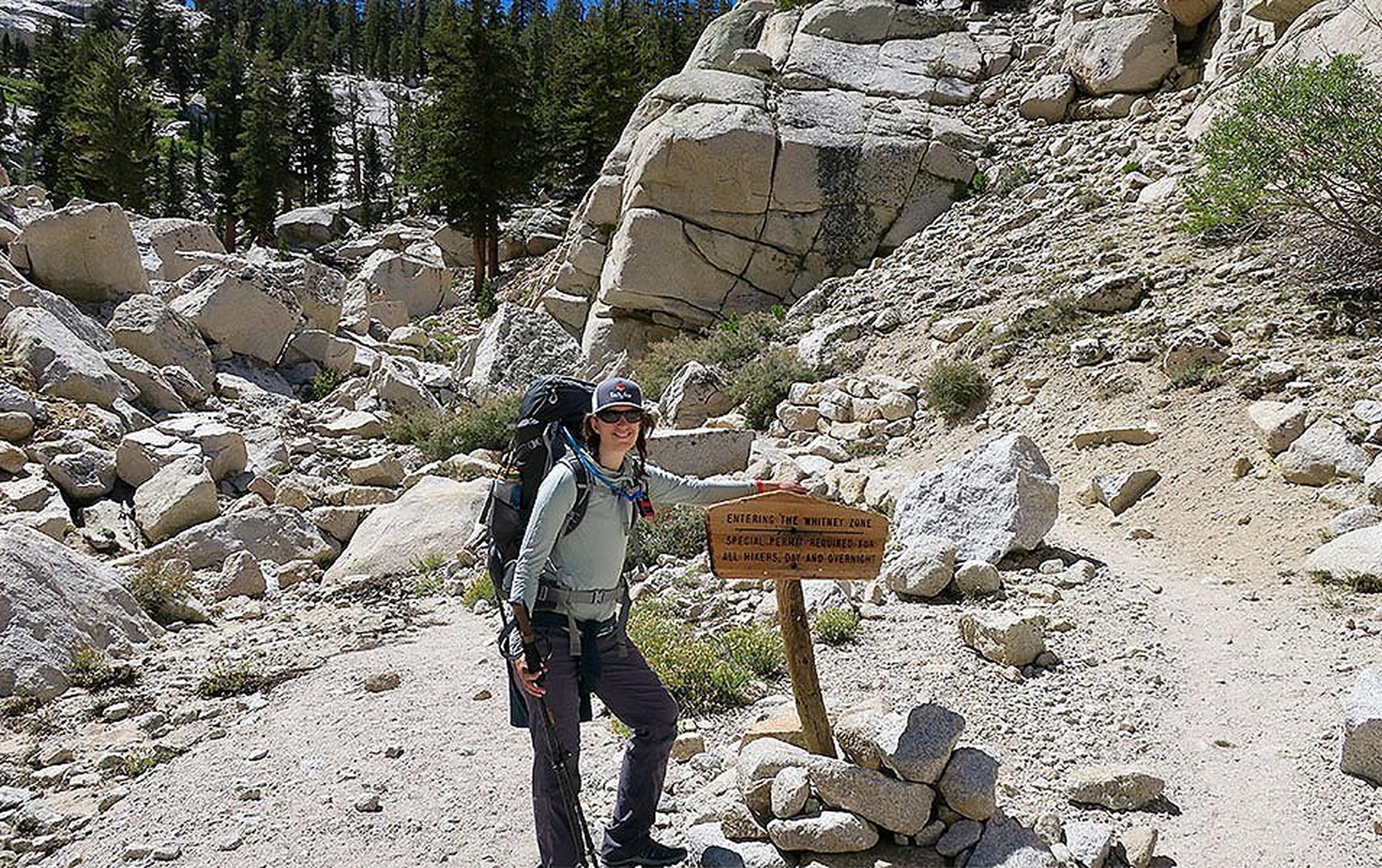 A woman stands at the base of the Mt. Whitney trail carrying a backpack and smiling for the camera.