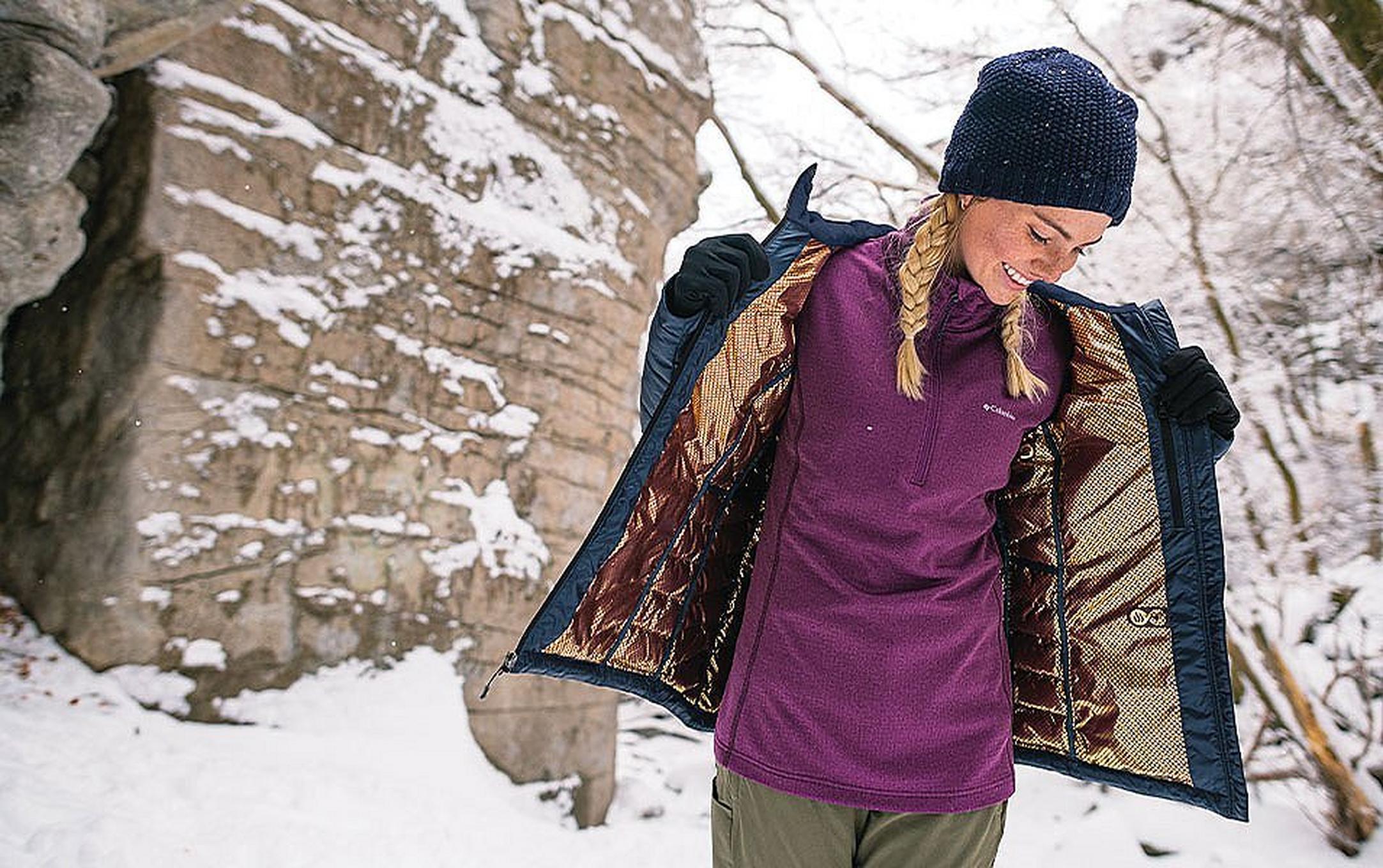 A woman standing near a snowy rock wall with her jacket open.