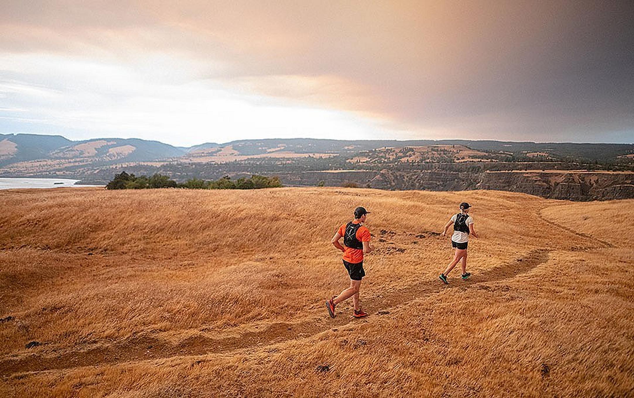 A man in a red shirt jogs behind a woman wearing white as they run along a narrow trail through an open field near a river.
