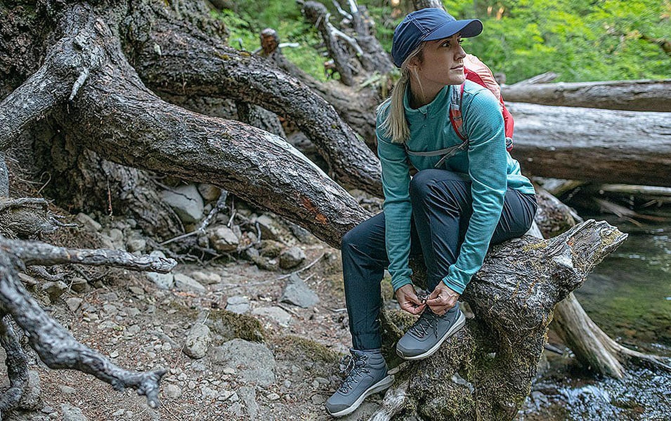 A woman wearing a blue Columbia Sportswear shirt sits on a log in the forest tying the laces of her hiking shoes.