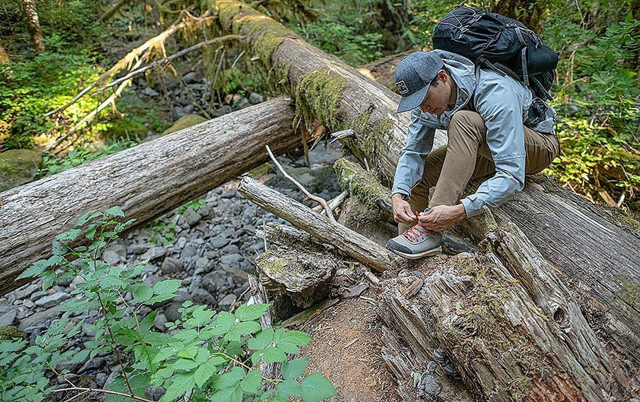 A man wearing a light blue Columbia Sportswear jacket stoops down on a mossy log in the woods to lace his hiking boots.