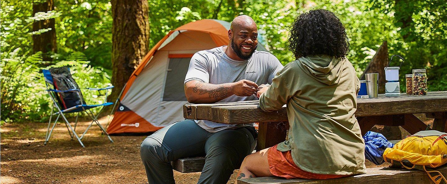 Two people sitting at a picnic table in woods in a campsite.
