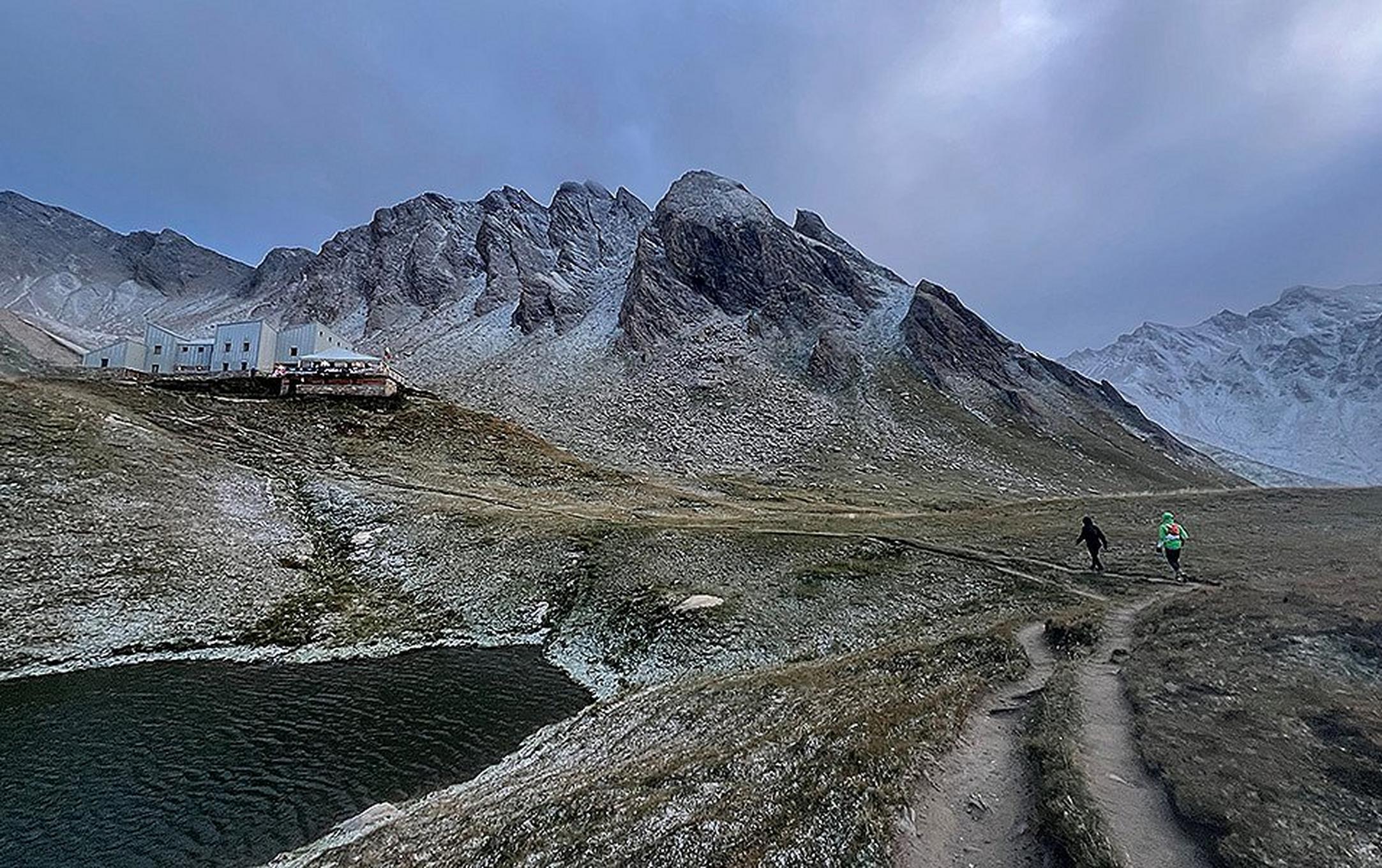 Two runners near a remote lodge built into the treeless alpine terrain above a lake. The runners are competing in the 200-mile Tor des Géants.