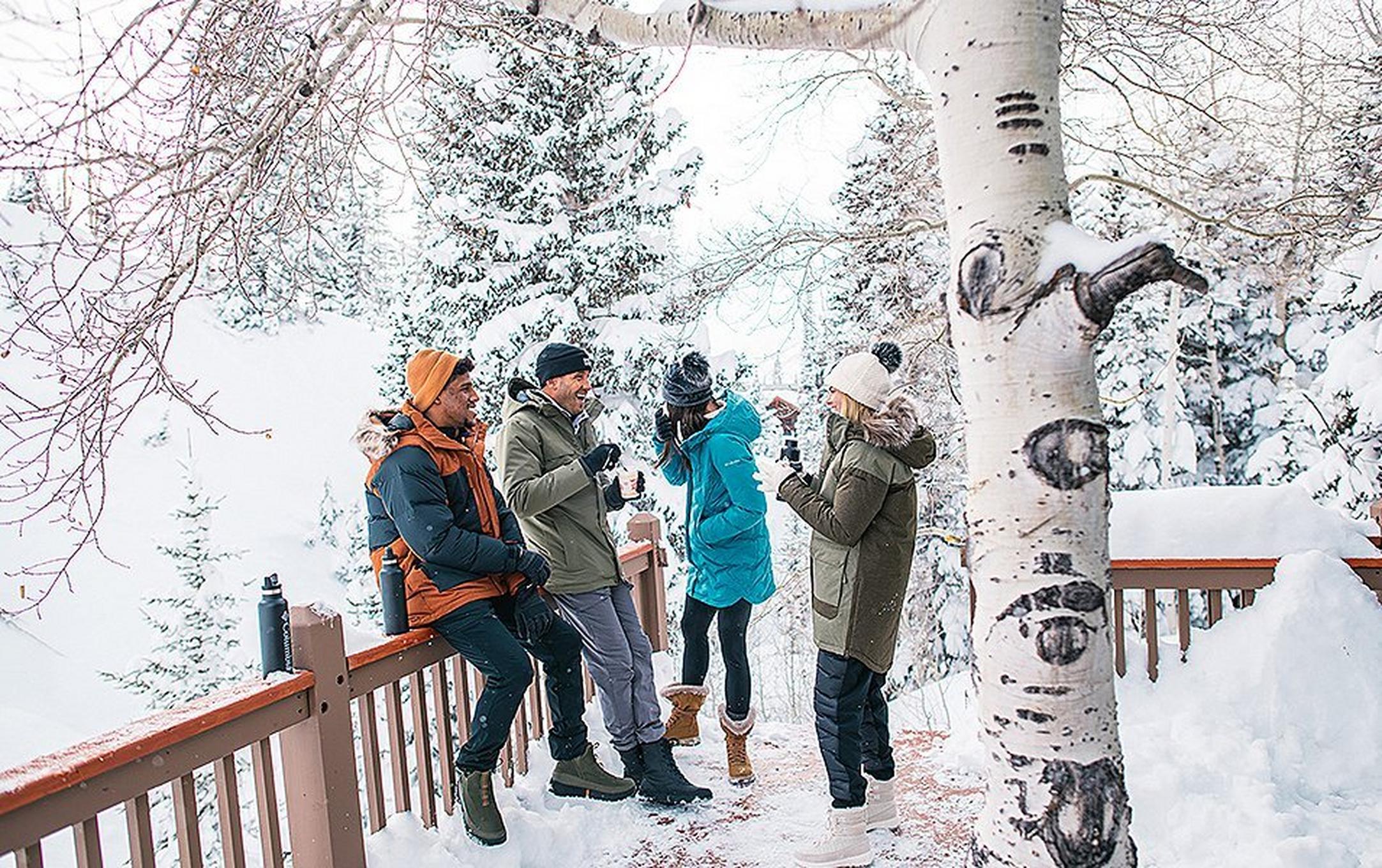 A group of friends stand outside a winter cabin drinking hot beverages and chatting.