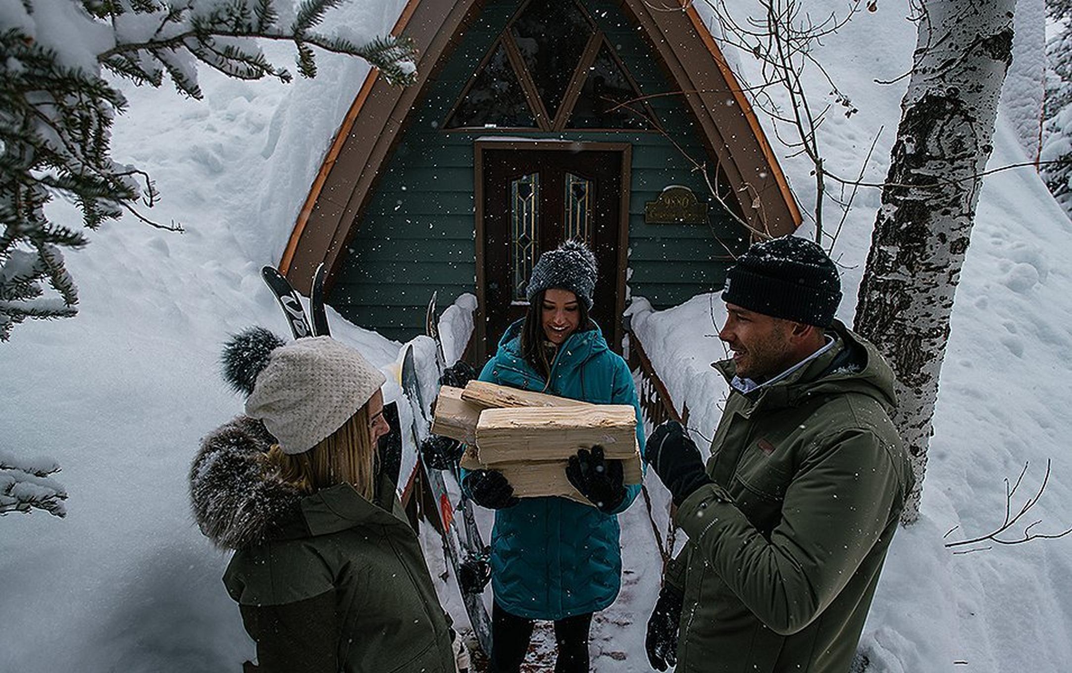 A group of friends stands outside a winter cabin chatting while one holds firewood.