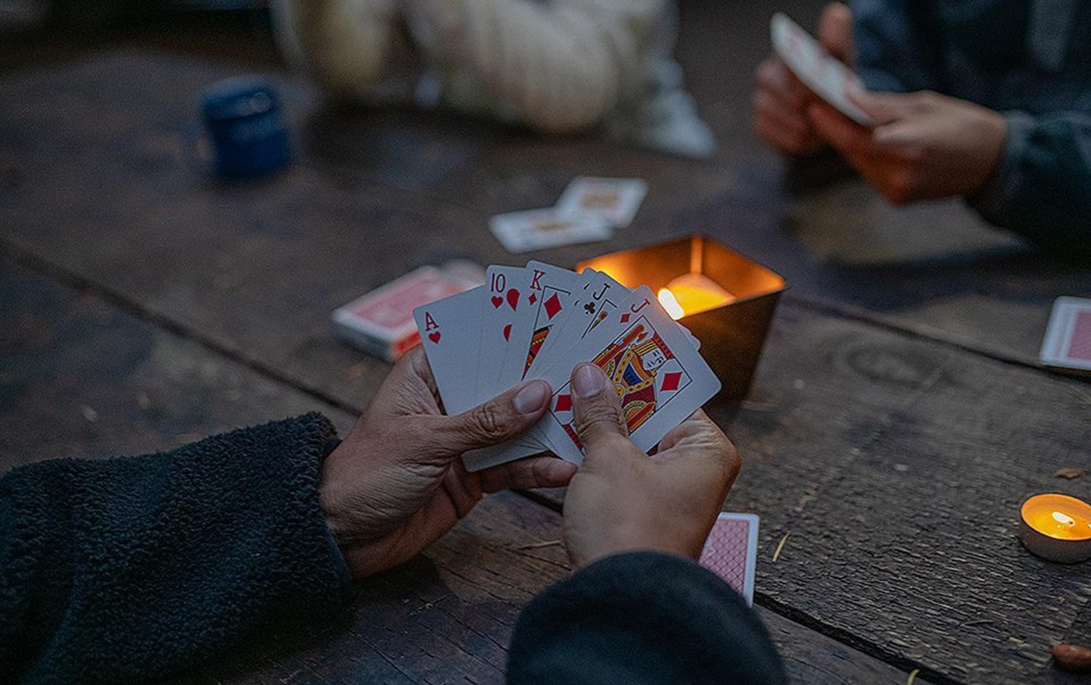 A close-up shot of someone’s hands holding playing cards at a rustic picnic table set with lighted candles.