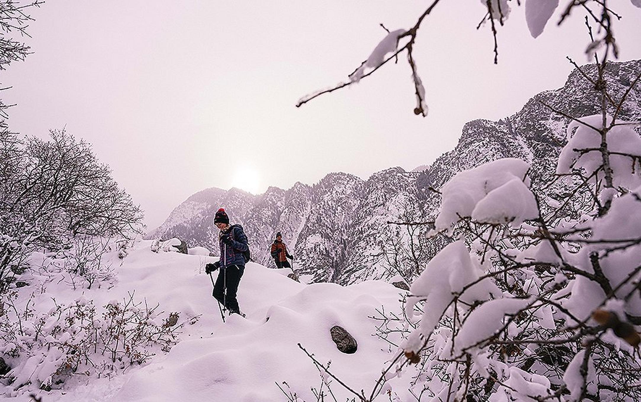 A woman treks through a snowy winter wonderland with another hiker and a stretch of mountains in the background.