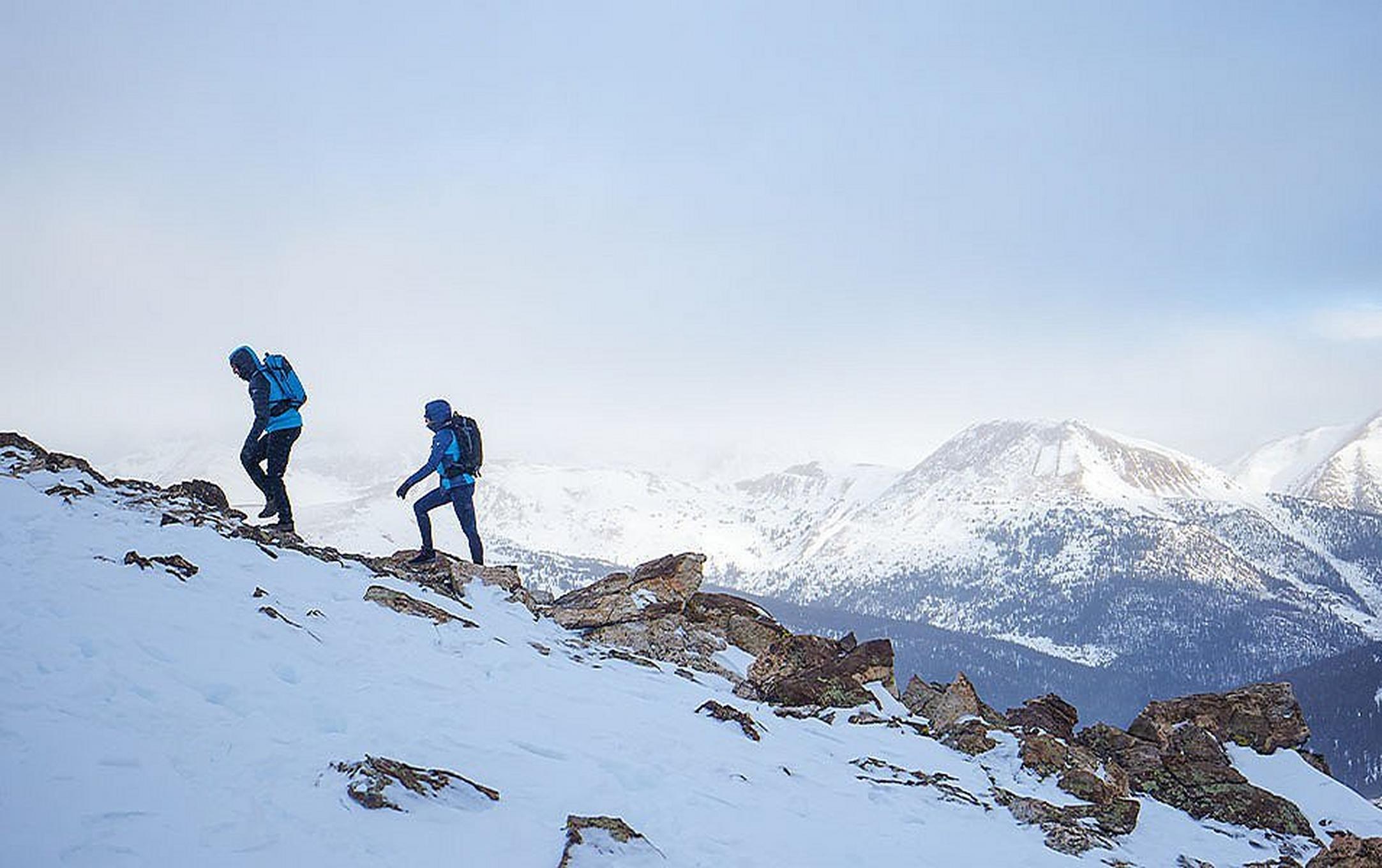 A pair of hikers wearing blue puffer jackets trek up a mountain trail in front of a scenic winter backdrop.