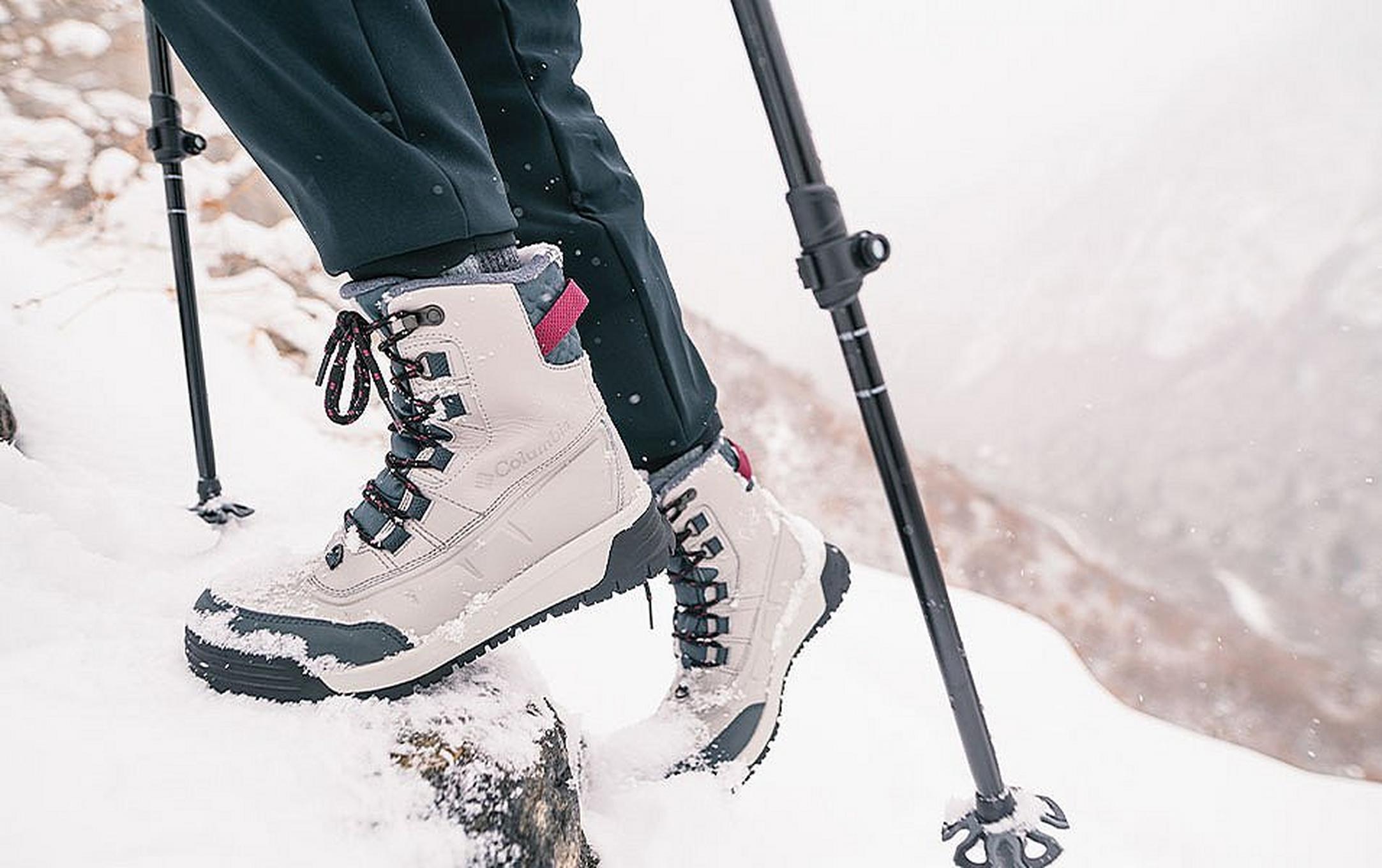 A close-up photo of a pair of white winter hiking boots and trekking poles as they climb up a snowy mountain trail.