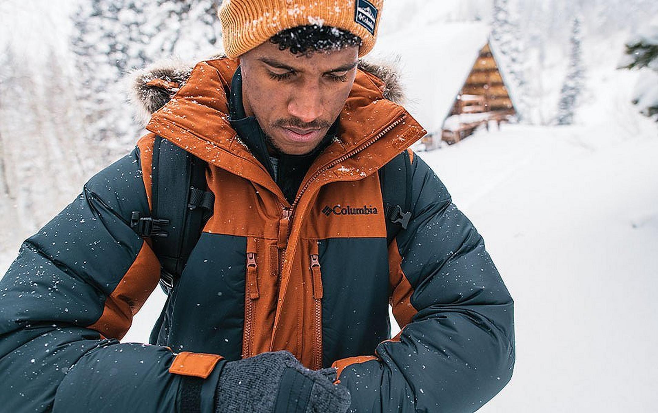 A man in an orange parka and beanie adjusts his gloves amid a scenic winter setting.