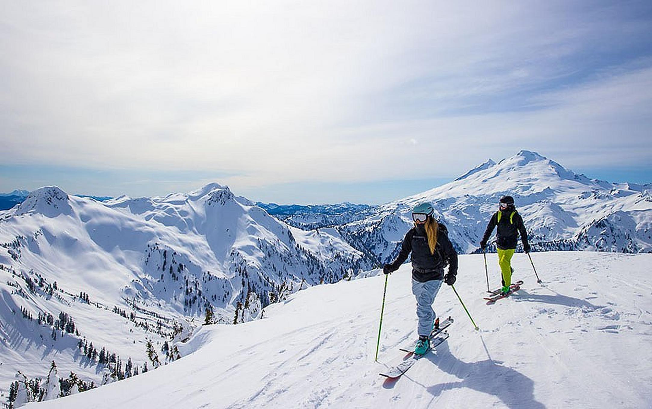 Two skiers stand on top of a gorgeous mountaintop on a sunny day with a sweeping panoramic view behind them.