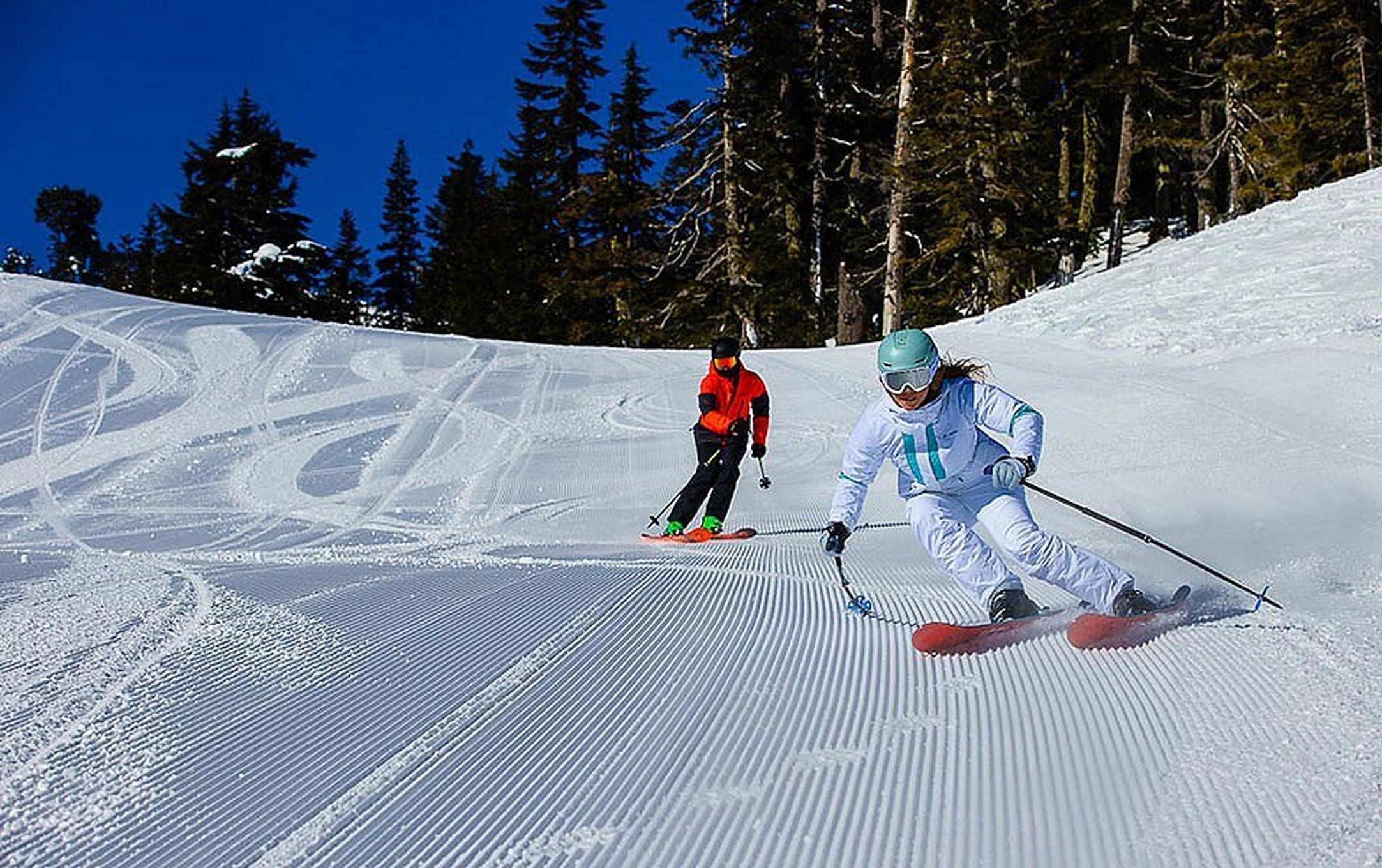 Two skiers rip down fresh corduroy tracks on a sunny bluebird day at the mountain.