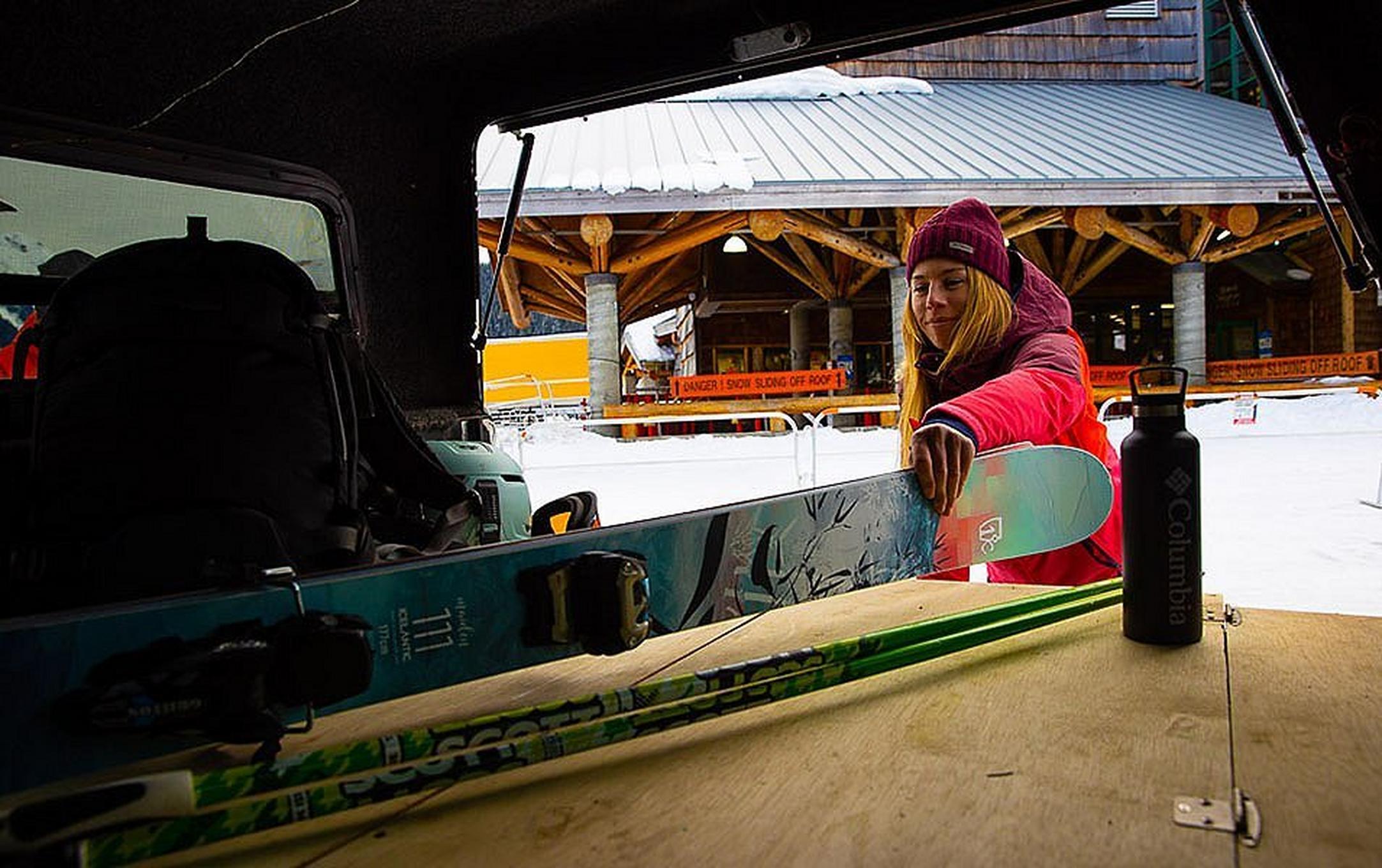 A woman in a reddish pin jacket pulls a set of skis out of her car with a ski lodge in the background.