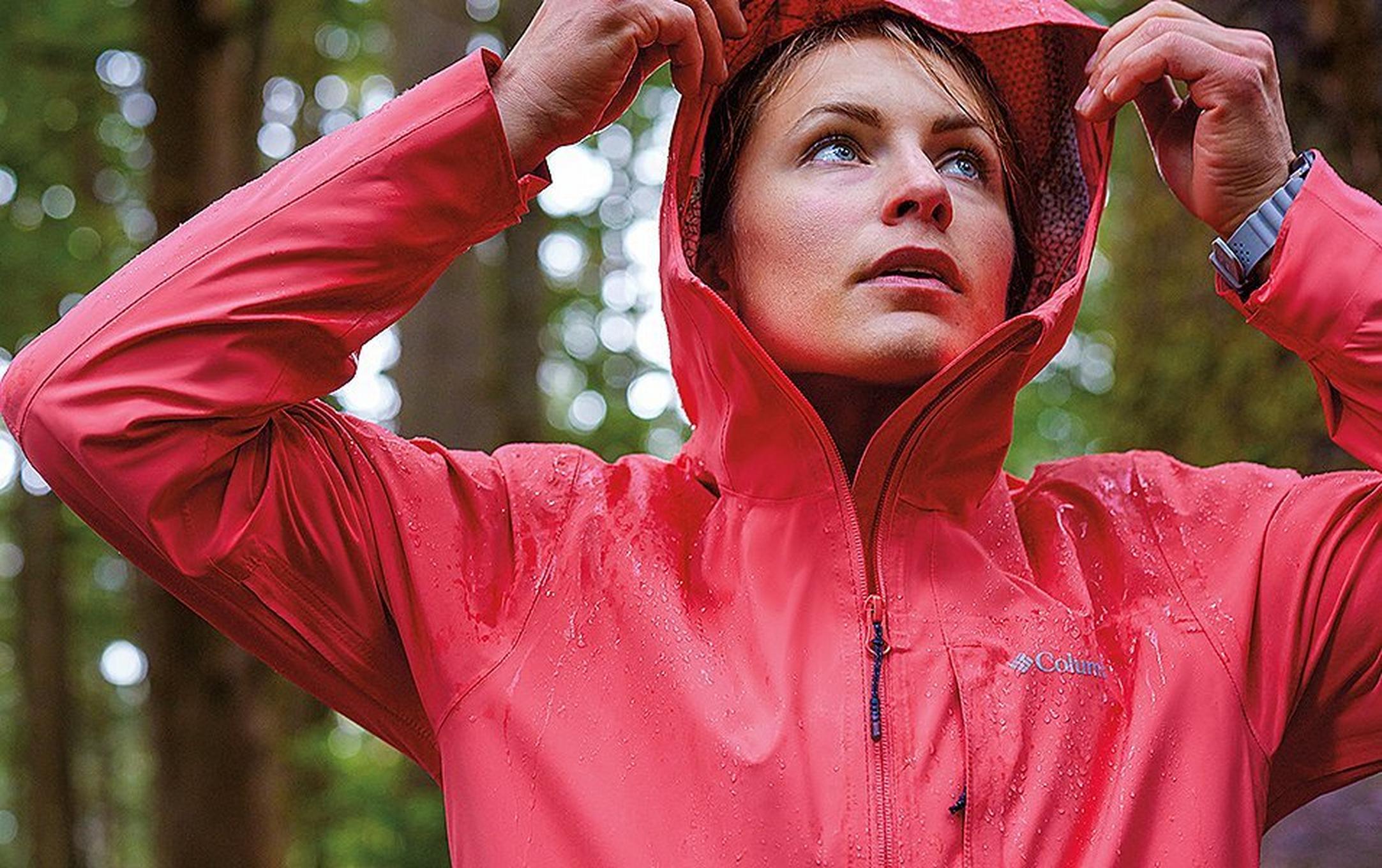 A woman wearing a red Columbia Sportswear Omni-Tech™ jacket pulls up her hood in a rainy forest with trees in the background.