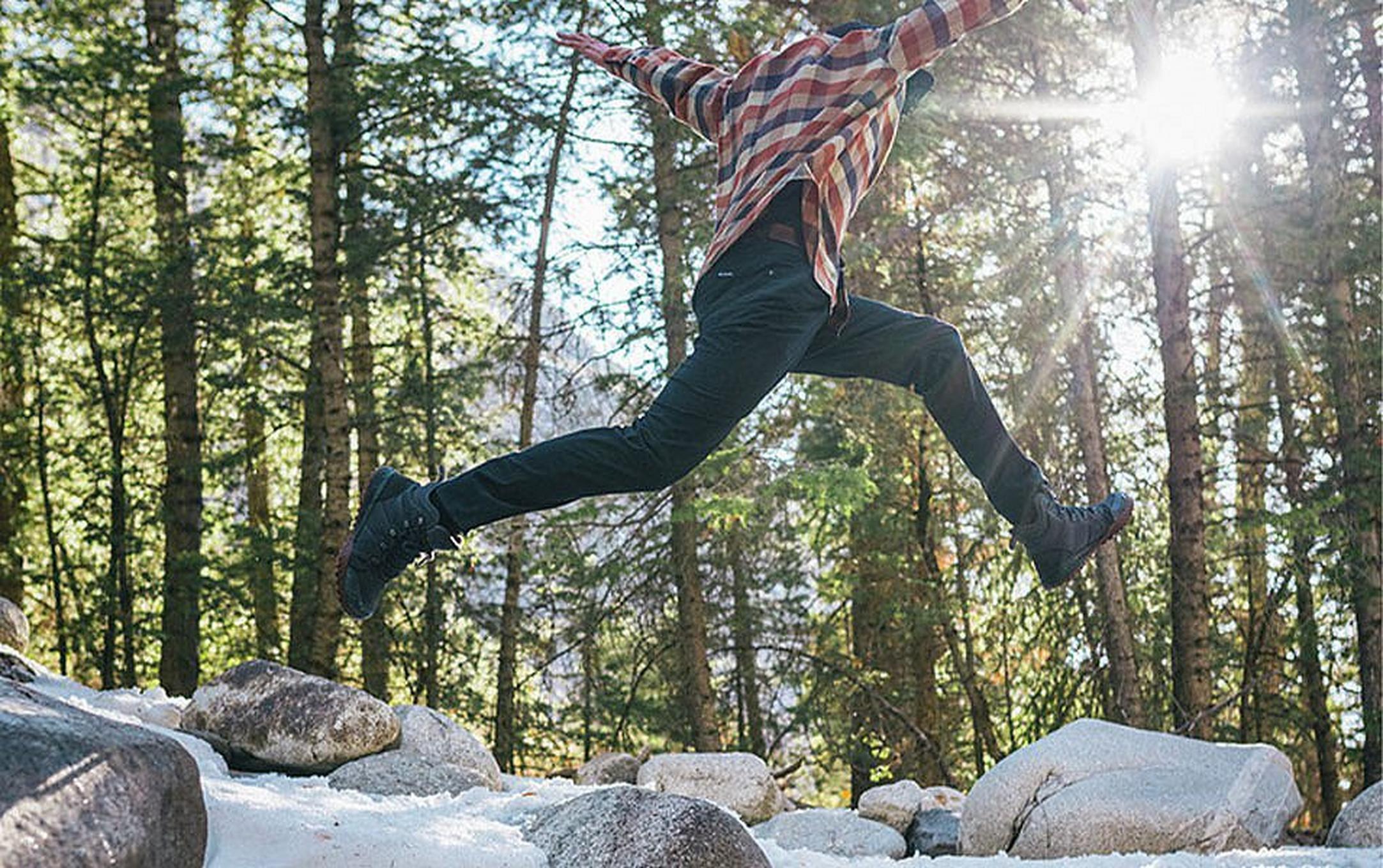 A man in a checkered red shirt and black pants leaps between two rocks with his arms in the air in the middle of a scenic wooded area.