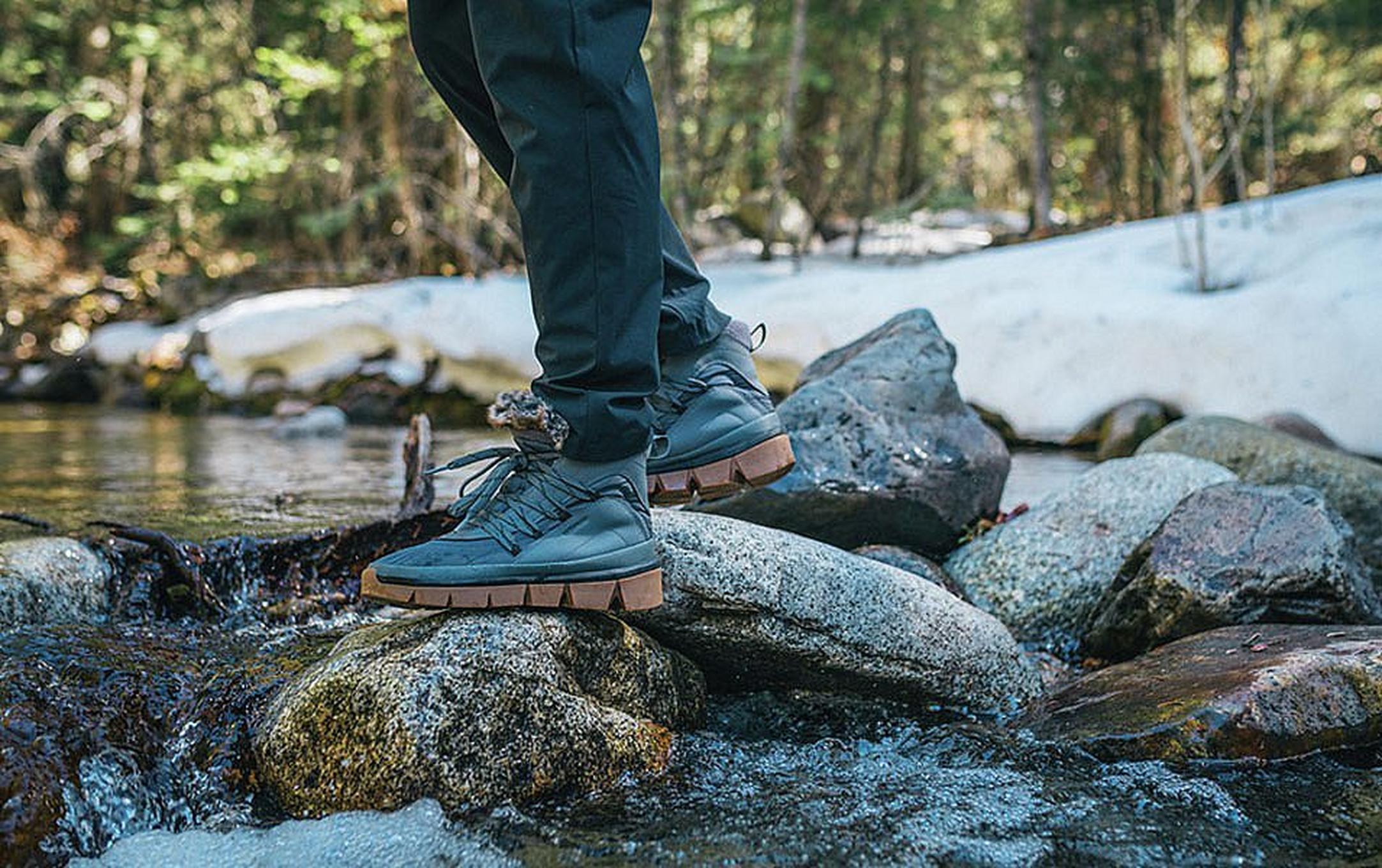 A waist-down photo of a man’s legs in black pants with dark boots stands on a cluster of mossy rocks in the middle of a creek.