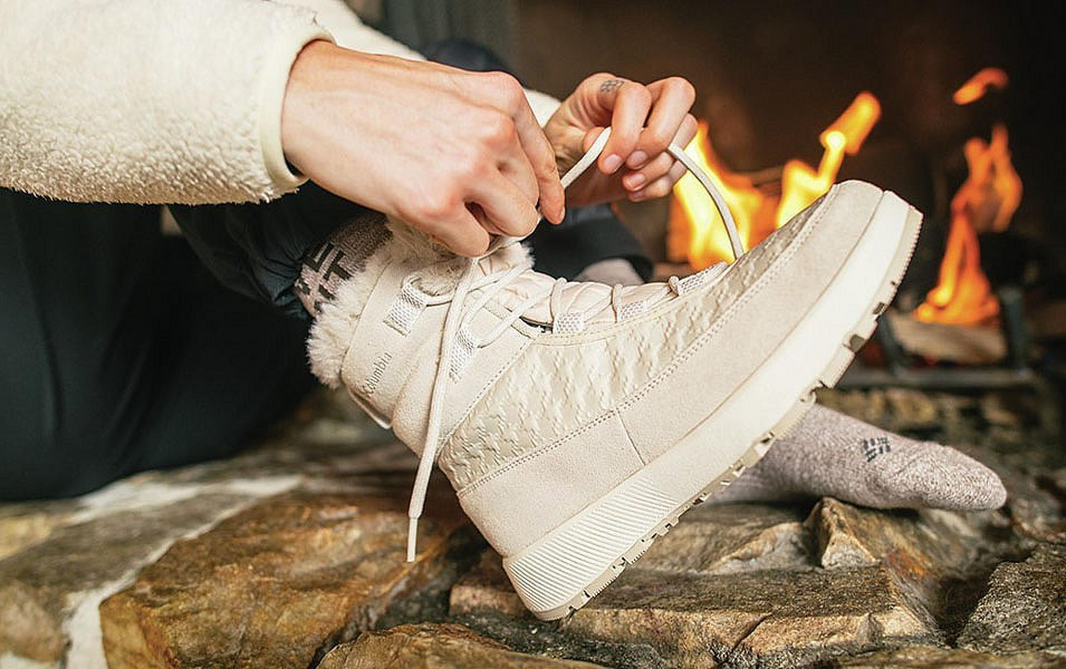 A close-up photo of a woman’s hands trying a pair of white Columbia Sportswear boots as a warmly lit fireplace glows in the background.