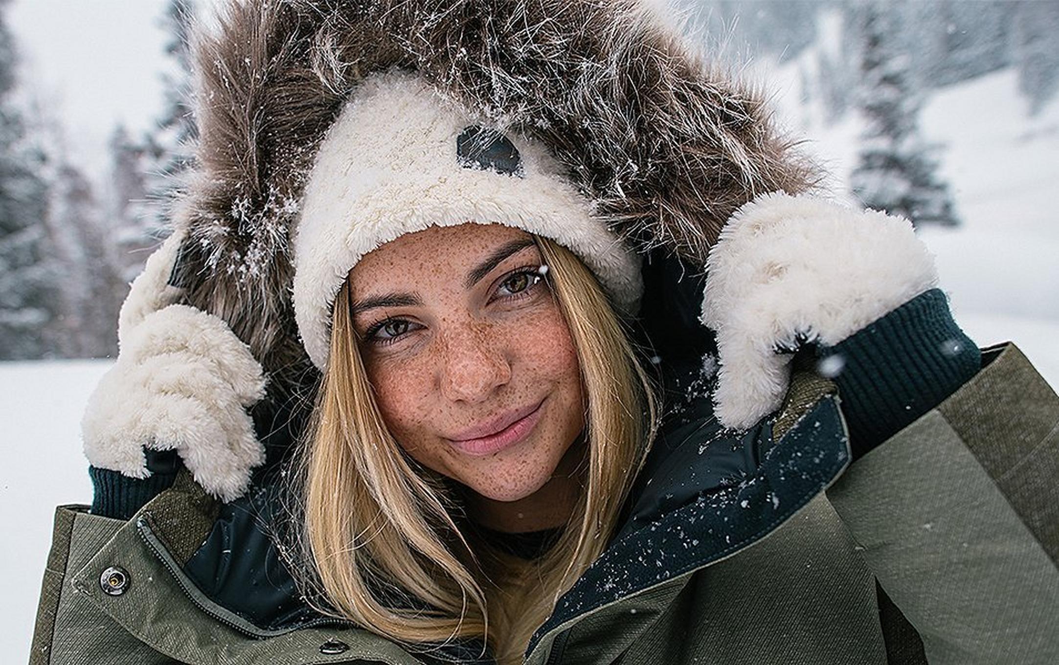 A woman stands in the snow wearing fluffy gloves and a jacket with a faux fur trim smiling at the camera.