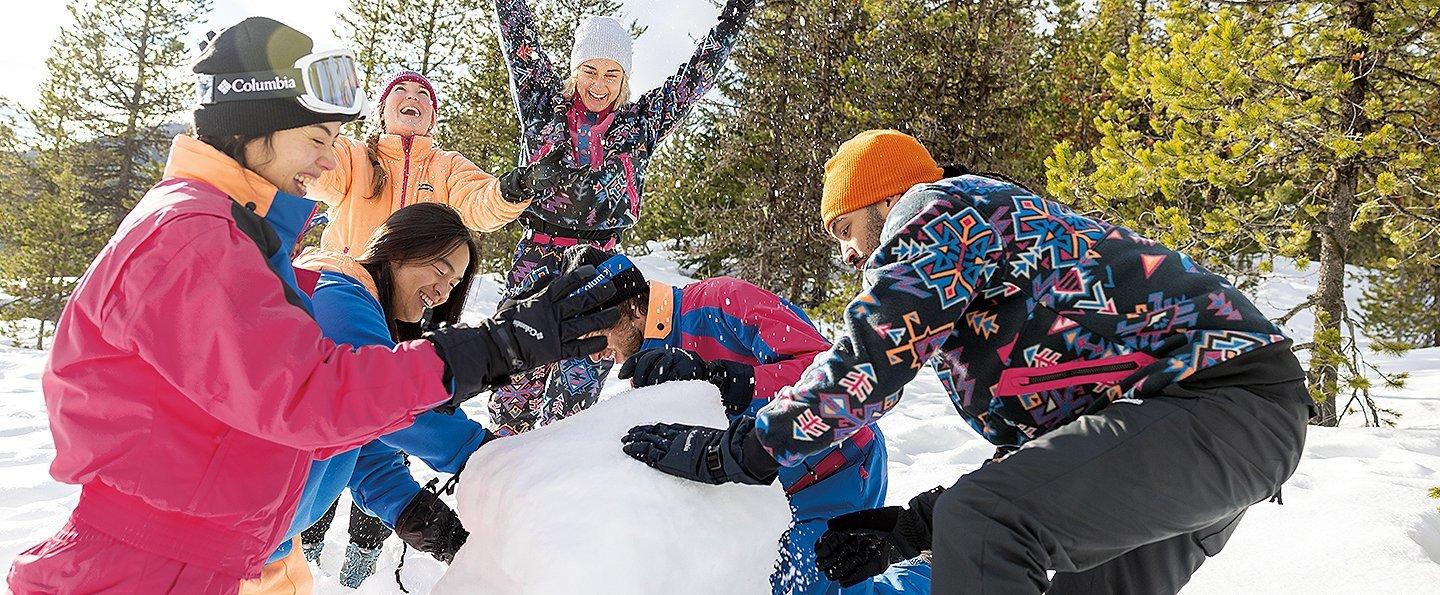 A group of skiers laughs together as they build a snowman.