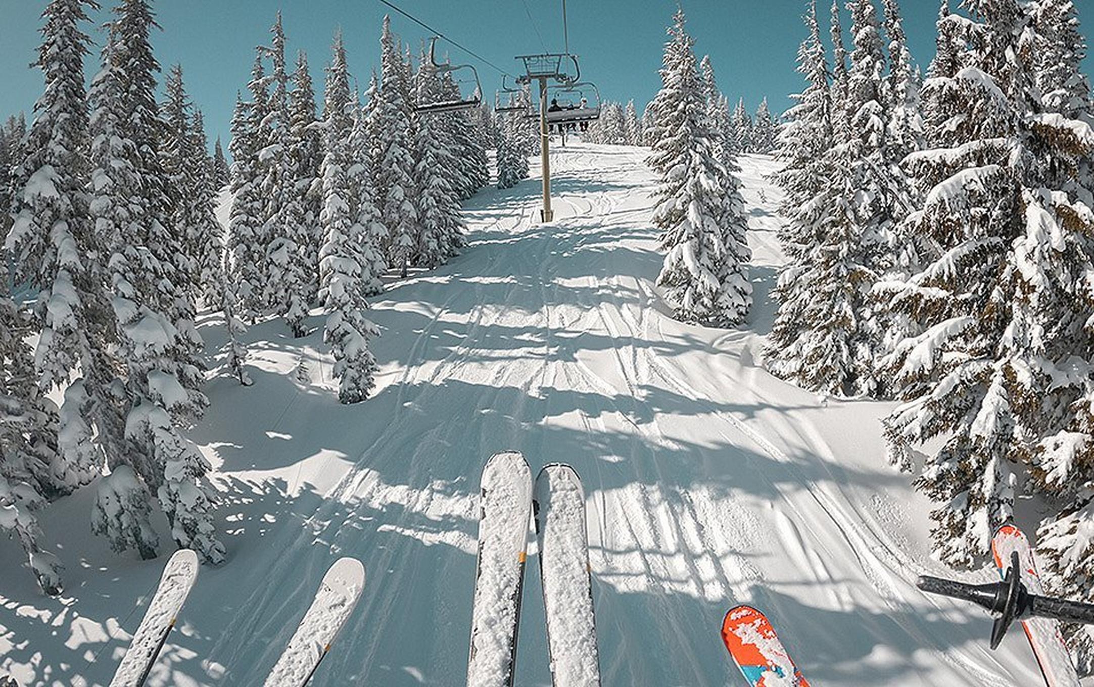 Three skiers are riding up a chairlift. Between their ski tips you can see a snow-laden slope with deep tracks.