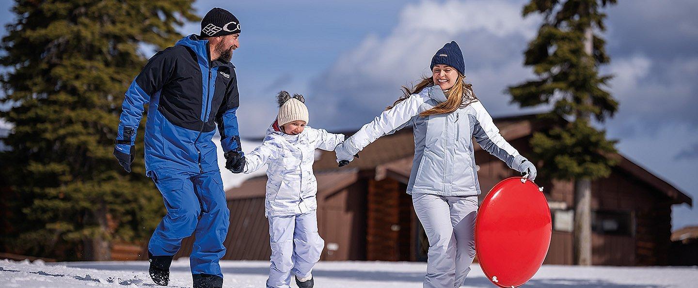 A man and woman swing a child between them, holding hands. The woman is carrying a round red sled.