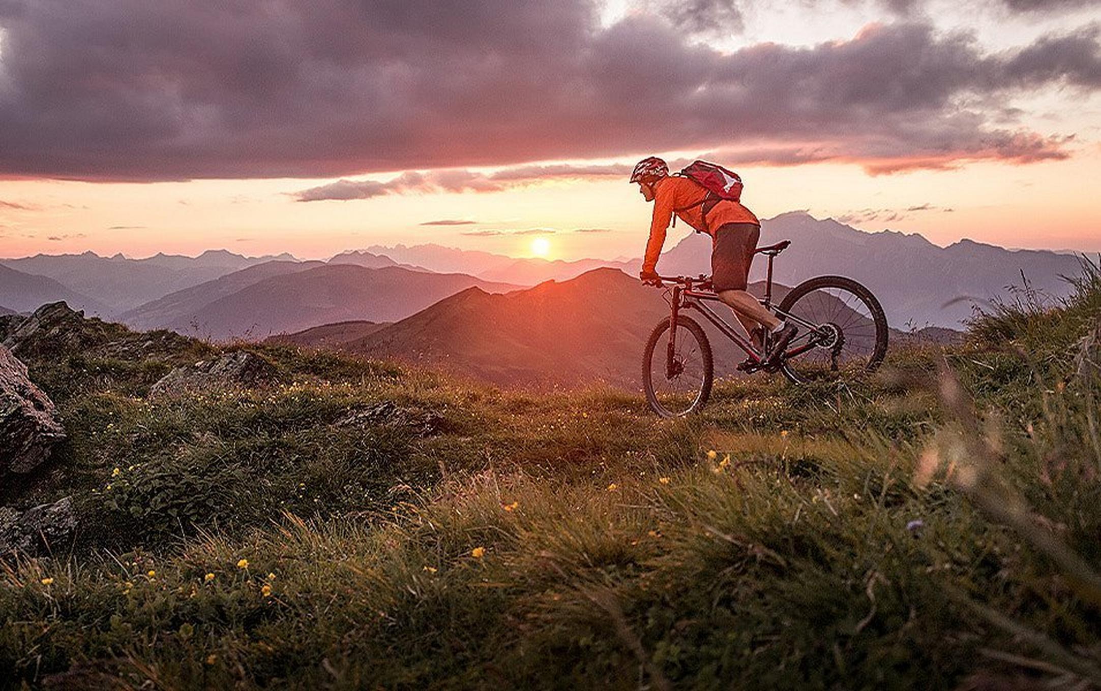 A man rides his mountain bike through a flower-strewn meadow, with jagged peaks in the distance and the sun setting behind them as clouds gather.