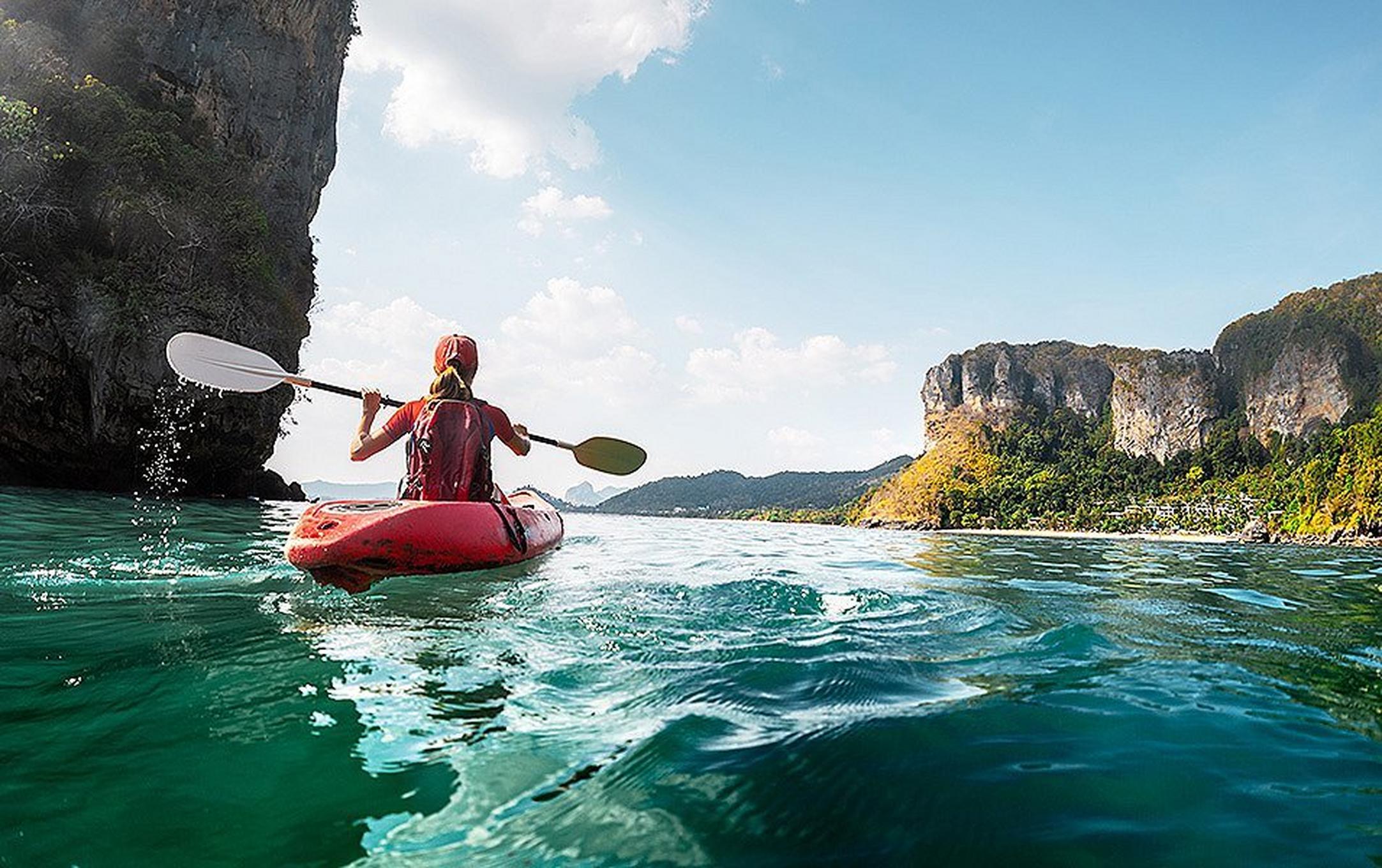 A woman in a red kayak paddles across sparkling blue water. There are steep cliffs of both sides.