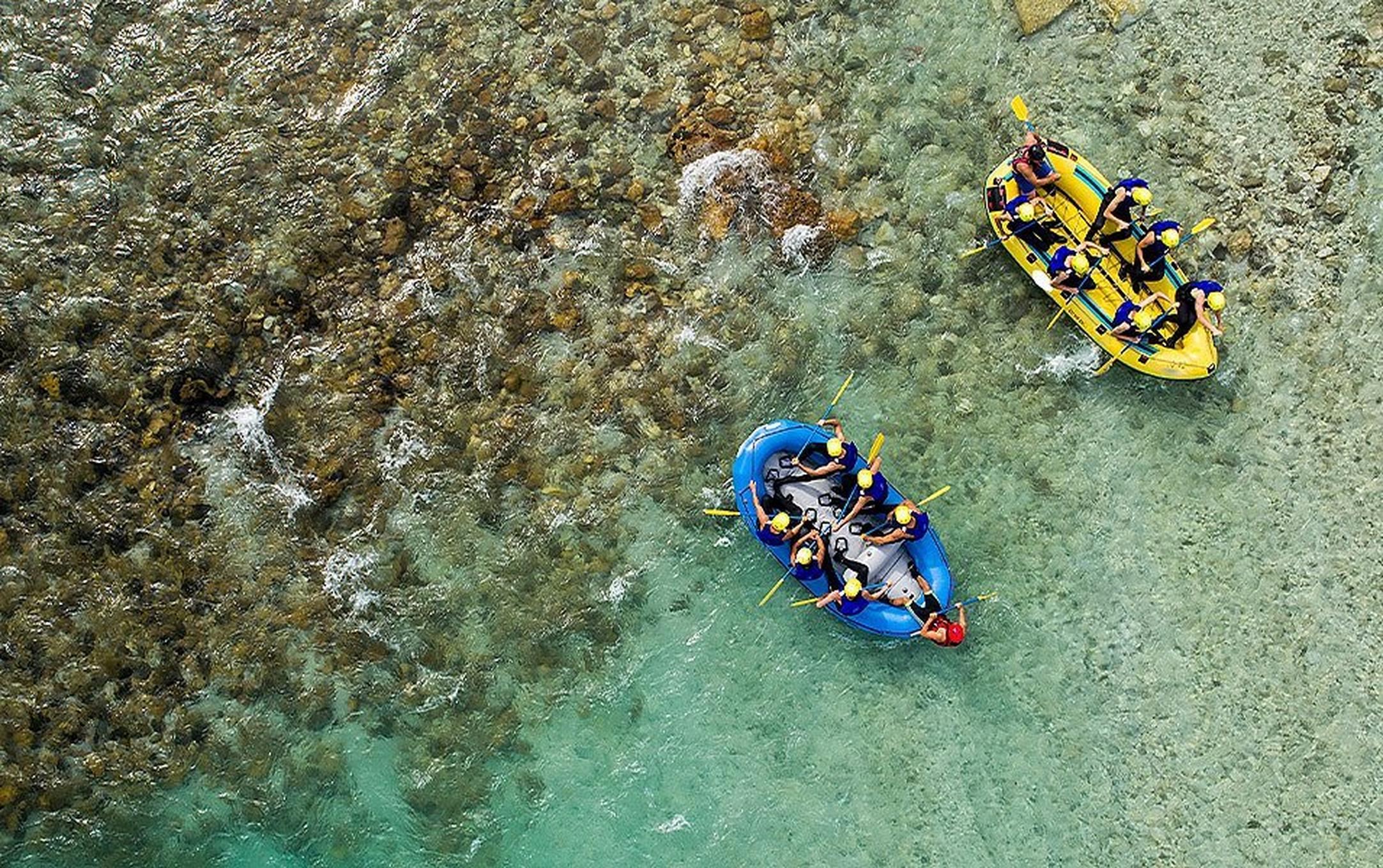 Two whitewater rafts, one yellow and the other blue, each carrying seven people, float side by side down a shallow stretch of river.
