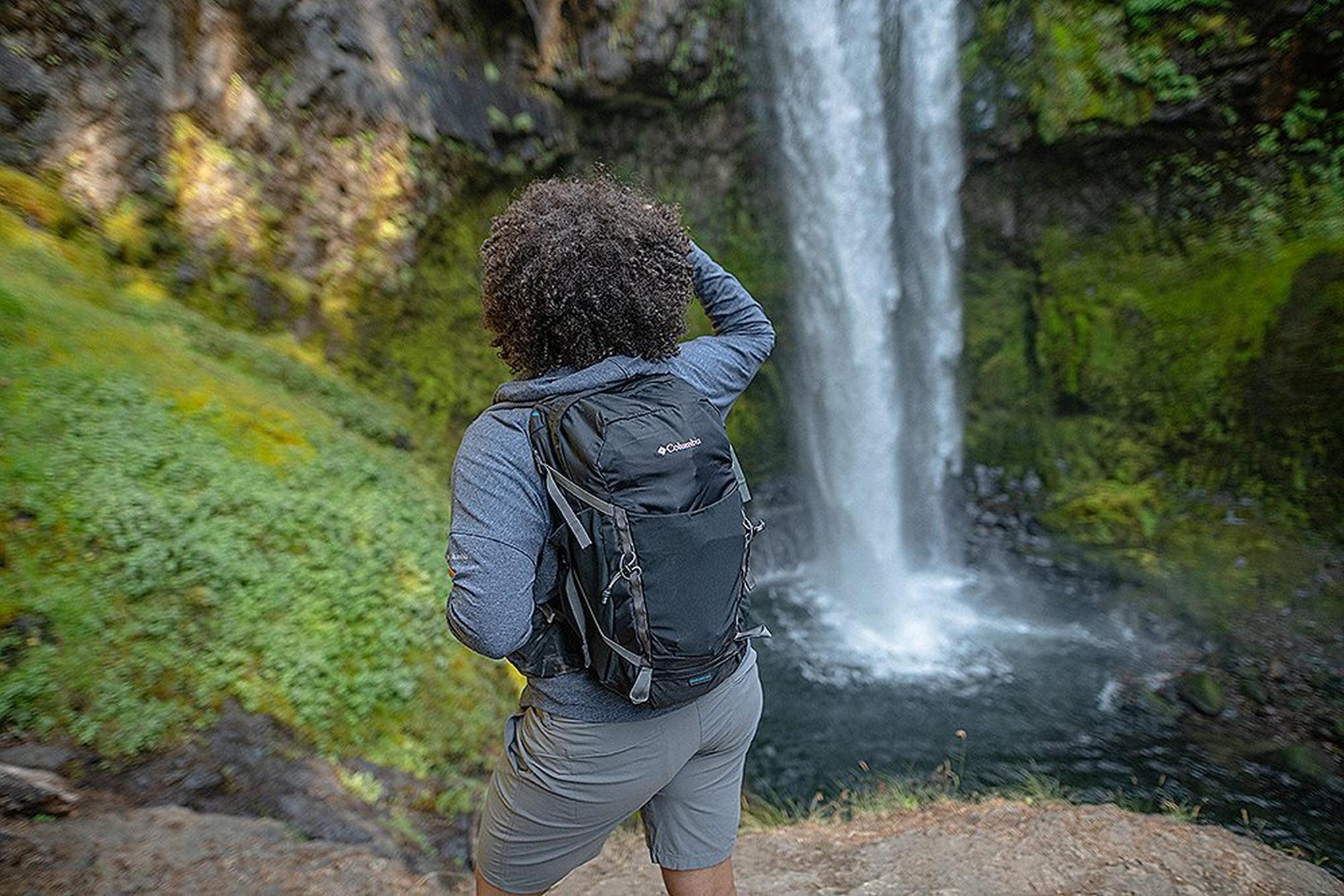 A backpacker pauses to look at a gorgeous cascading waterfall in the middle of a lush green forest.