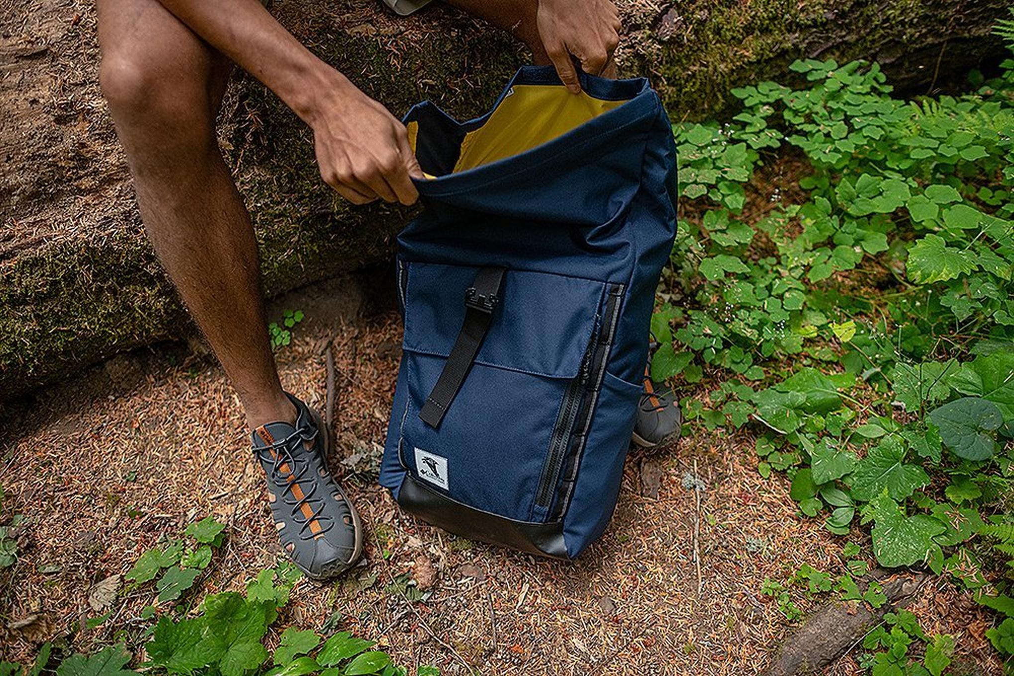 A close-up shot of a backpack on the ground in a scenic forest as a hiker’s hand digs through it.