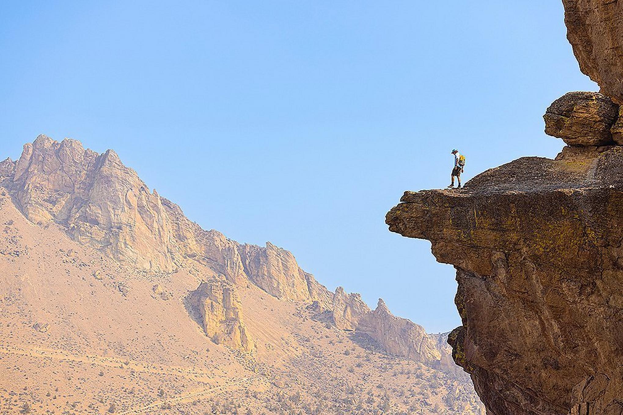 A backpacker stands on a rocky cliff in the distance staring out over a beautiful canyon.
