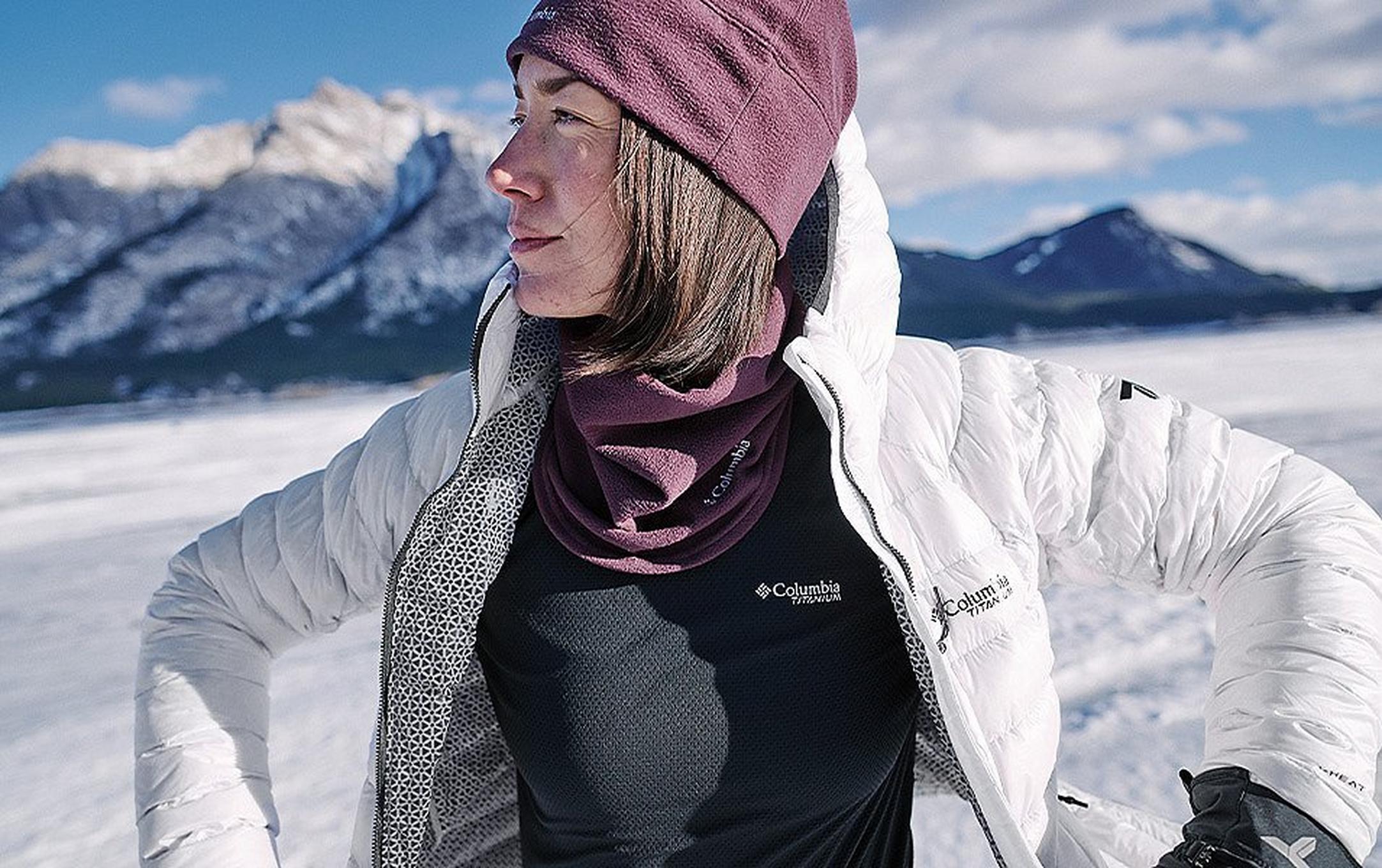 A woman wearing a white Columbia Sportswear Titanium Jacket, a maroon fleece hat and neck gaiter, with snow-covered mountains in the background.