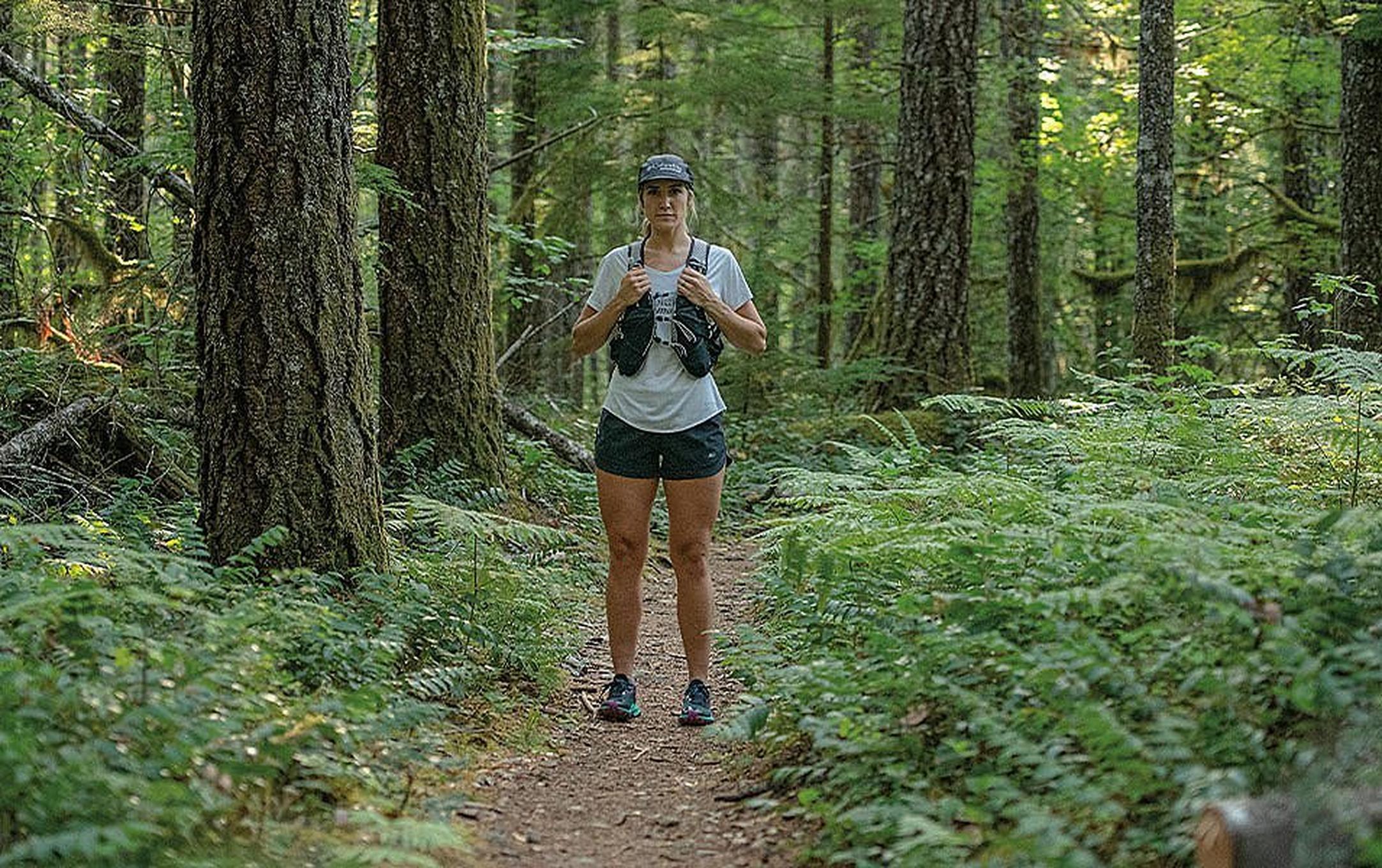 A woman in trail running gear stands in a forest staring at the camera with a bold stance.