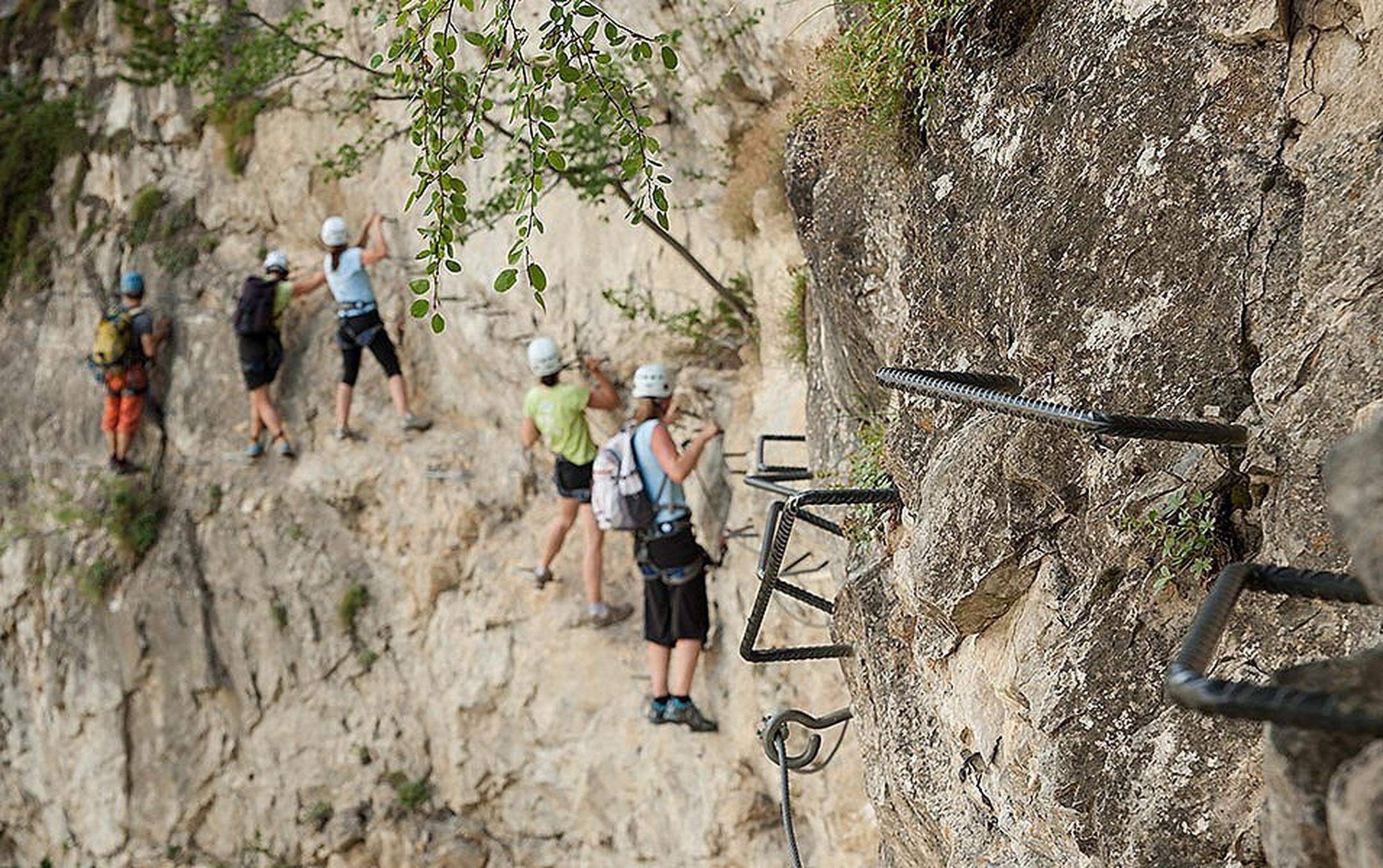 Five people appear to be climbing side by side on a steep rock face but closer inspection reveals that they are using metal hand and footholds and a safety cable that creates an easy path along the cliff.